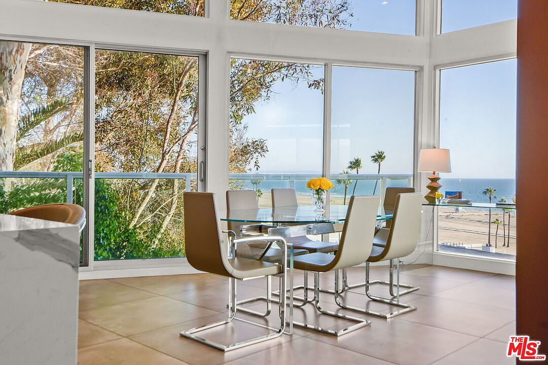 This bright dining area features a glass-topped table surrounded by four modern, light-colored chairs with sleek metal frames. The room is defined by expansive, floor-to-ceiling windows that provide an unobstructed, panoramic view of the beach and ocean beyond. The neutral tile flooring and minimalist decor create an airy, sophisticated atmosphere that emphasizes the coastal setting.