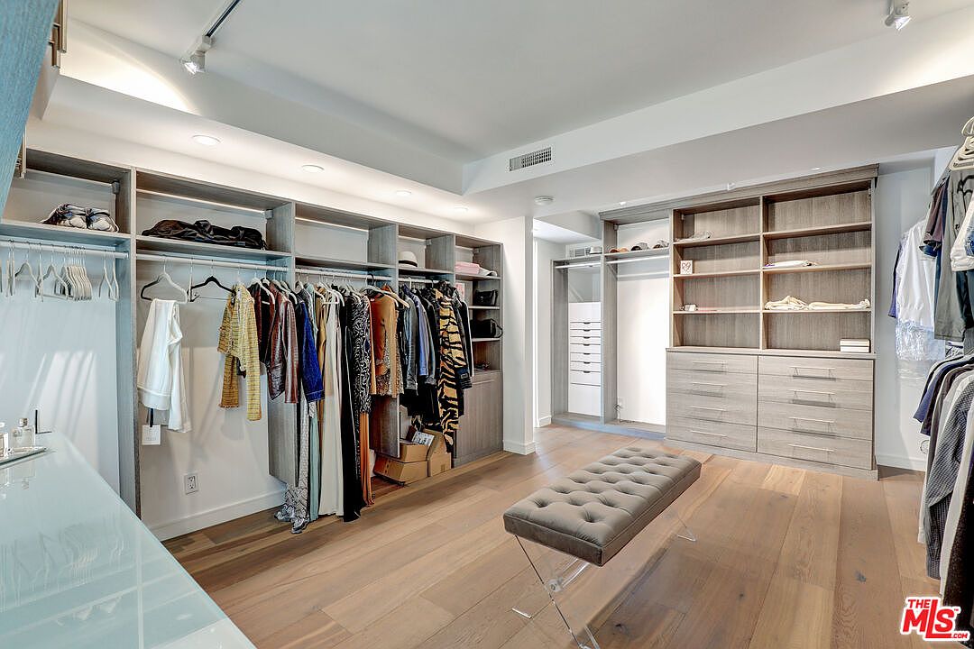 This spacious walk-in closet features extensive custom light wood cabinetry with a mix of hanging rods, open shelving, and a bank of drawers. A tufted grey ottoman sits in the center of the room on wide-plank wood flooring, providing a functional focal point. The space is brightly lit by recessed ceiling lights and track lighting, creating a clean and organized atmosphere.