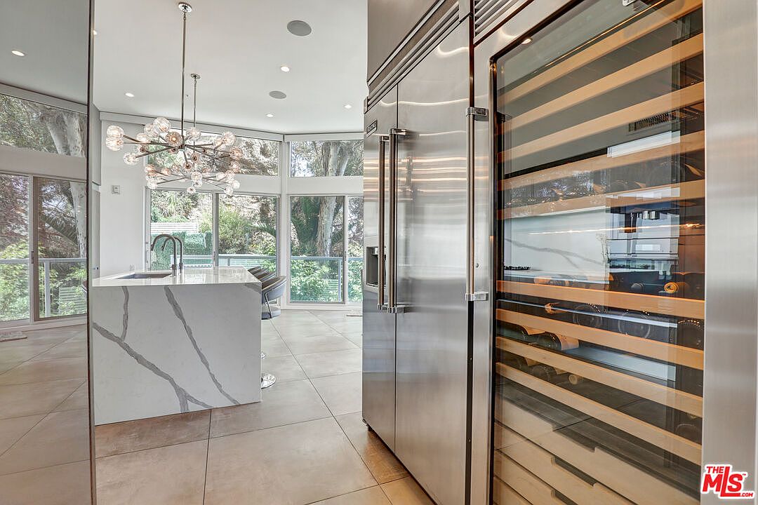 This modern kitchen features a large island with a stone countertop and a waterfall edge, complemented by a stainless steel refrigerator and a built-in wine cooler. The space is illuminated by a striking, multi-bulb chandelier and benefits from natural light streaming through large windows that overlook the surrounding greenery. The neutral-toned tile flooring and sleek cabinetry create a sophisticated, contemporary atmosphere.