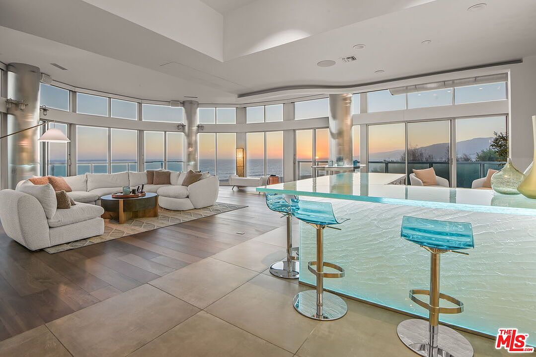 This expansive open-concept living area features a modern aesthetic with a large, curved sectional sofa and a central coffee table set on a patterned area rug. A prominent kitchen island with a translucent, illuminated base and bar seating anchors the foreground, while floor-to-ceiling windows provide panoramic ocean views at sunset. The space is defined by its seamless transition between the living and kitchen zones, highlighted by sleek, light-toned wood flooring and a minimalist architectural design.