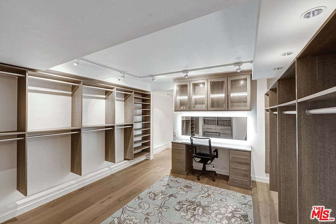 This spacious walk-in closet features extensive light wood cabinetry with open shelving and hanging rods lining the walls. A central vanity area includes a mirror, integrated lighting, and a desk space with a black office chair, all set against a light-toned wood floor covered by a patterned area rug. The room is illuminated by recessed ceiling lights and track lighting, creating a bright and organized dressing environment.