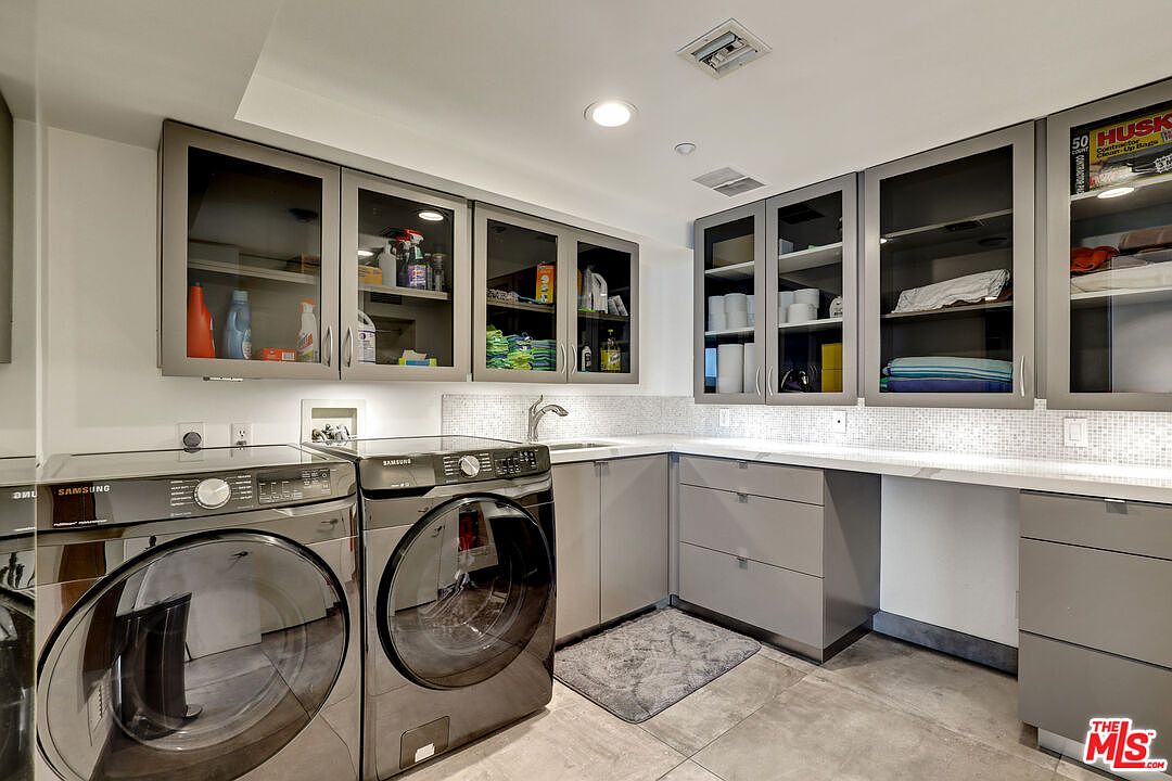 This modern laundry room features sleek, dark-toned front-loading Samsung washer and dryer units positioned beneath a long, light-colored countertop. Above the workspace, grey cabinetry with glass-front doors provides ample storage, complemented by a tiled backsplash and a built-in sink. The space is well-lit with recessed lighting and finished with neutral-toned floor tiles, creating a clean and functional utility area.
