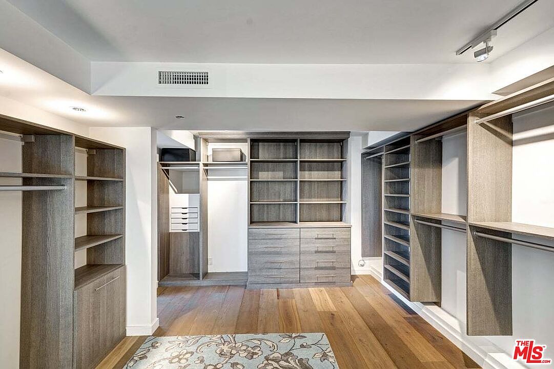 This spacious walk-in closet features custom light-toned wood cabinetry with a mix of open shelving, hanging rods, and built-in drawers. The room is finished with warm-toned wood flooring and a patterned area rug, creating a clean and organized aesthetic. The perspective is a wide-angle view looking into the center of the closet, highlighting the extensive storage capacity.