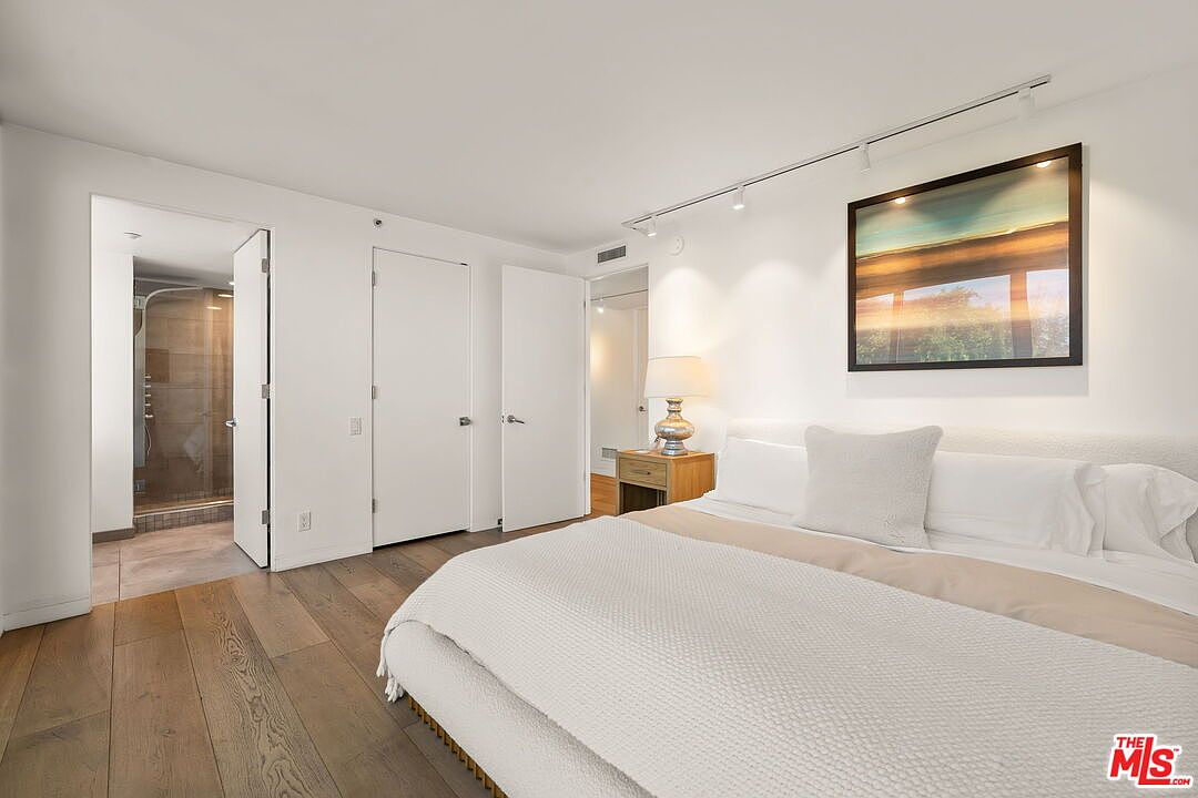 This bright and minimalist primary bedroom features light wood flooring and crisp white walls, creating an airy and serene atmosphere. A large, plush bed with neutral bedding serves as the focal point, complemented by a bedside table with a lamp and a framed landscape artwork above the headboard. The room includes a direct view into an attached bathroom with a glass-enclosed shower, emphasizing a clean, modern design aesthetic.