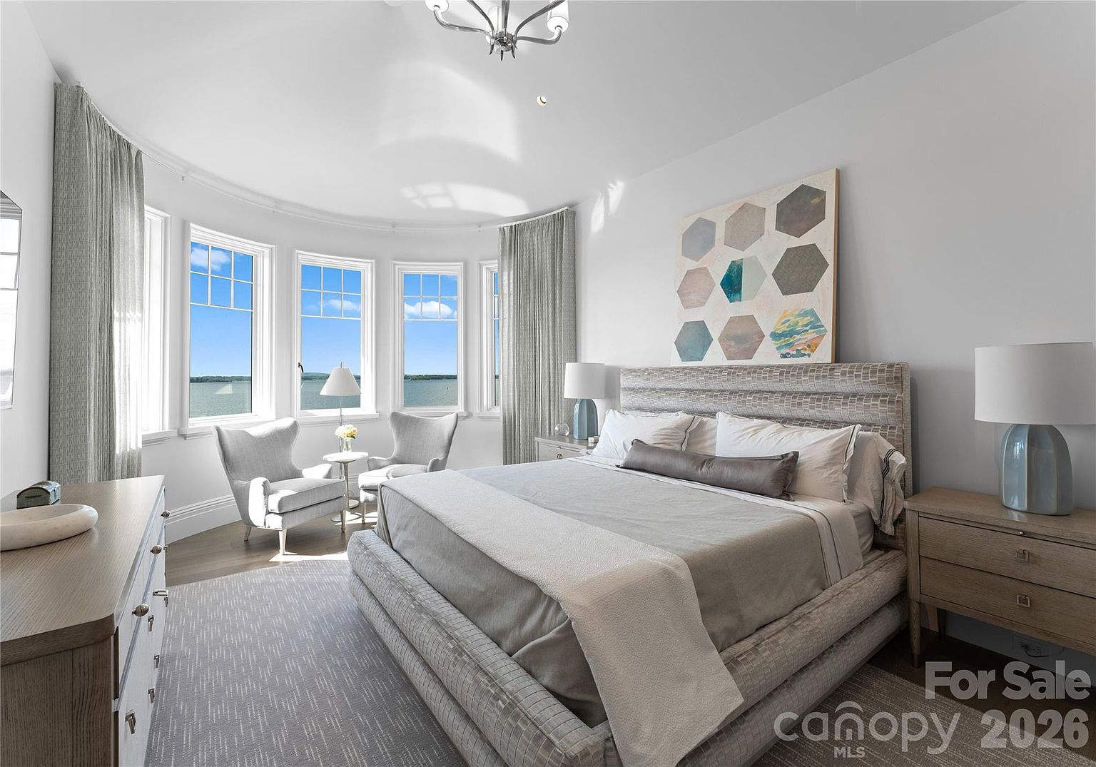 This primary bedroom features a neutral, sophisticated color palette with a large upholstered bed as the focal point. A curved wall of windows provides expansive water views and frames a cozy seating area with two armchairs and a small table. The room is accented by a geometric art piece above the headboard and matching bedside lamps, creating a serene and luxurious atmosphere.