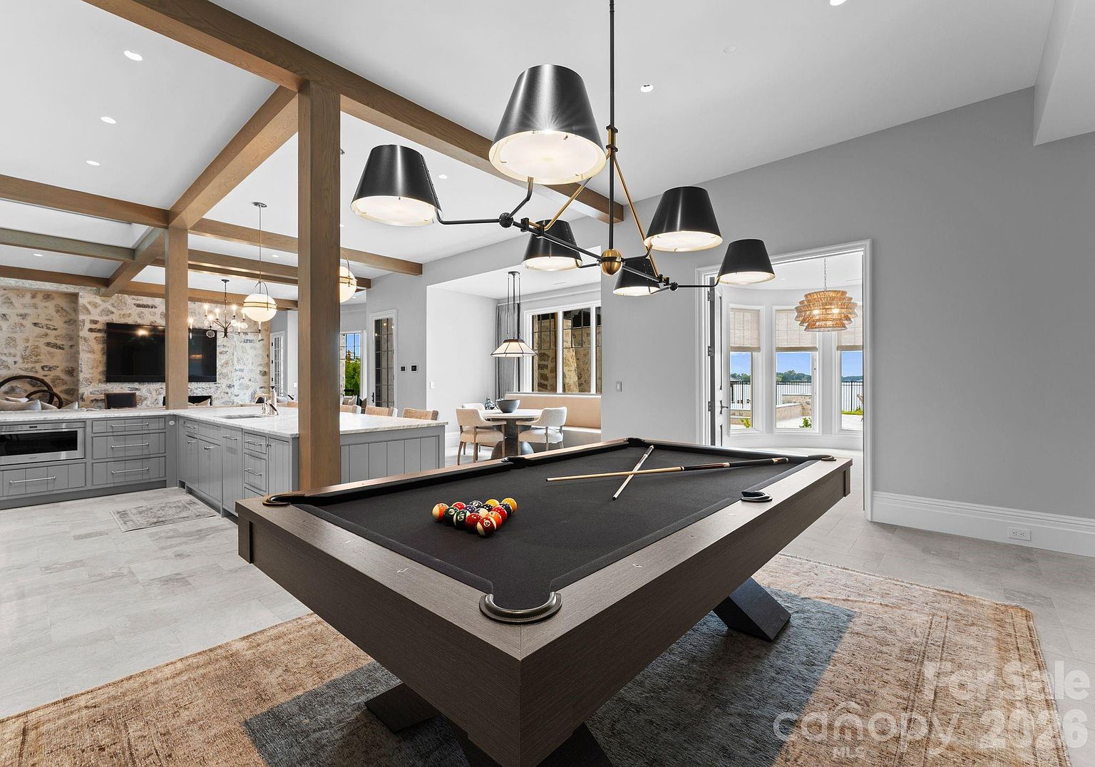 This open-concept living space features a prominent pool table as the central focal point, set beneath a large, multi-armed modern chandelier. To the left, a spacious kitchen with light-colored cabinetry and a stone-clad wall is visible, while a dining nook with a built-in bench sits in the background. The room flows seamlessly toward a set of glass doors that open to an outdoor view, creating a bright and airy atmosphere.