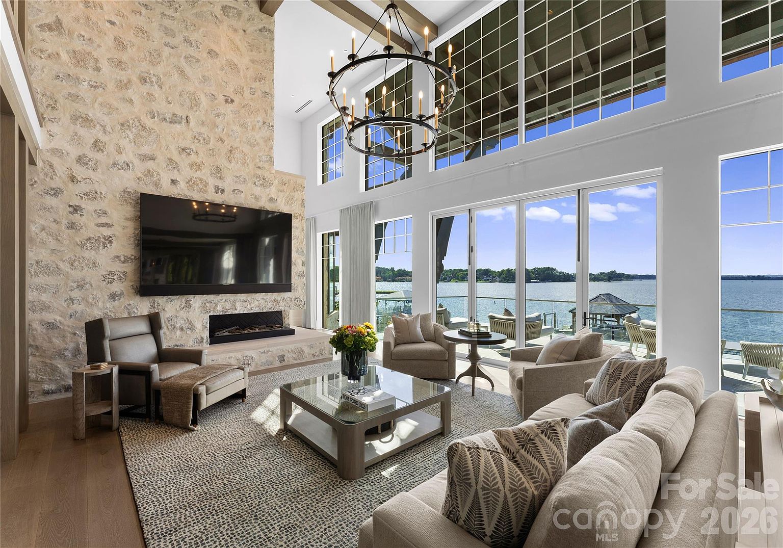 This expansive living room features a dramatic stone-clad chimney breast that serves as a focal point, complemented by a large television mounted above a linear fireplace. The space is bathed in natural light from tall windows that offer panoramic views of the lake, creating an airy and sophisticated atmosphere. A neutral-toned sectional sofa, a glass-topped coffee table, and a large iron chandelier complete the refined, open-concept design.