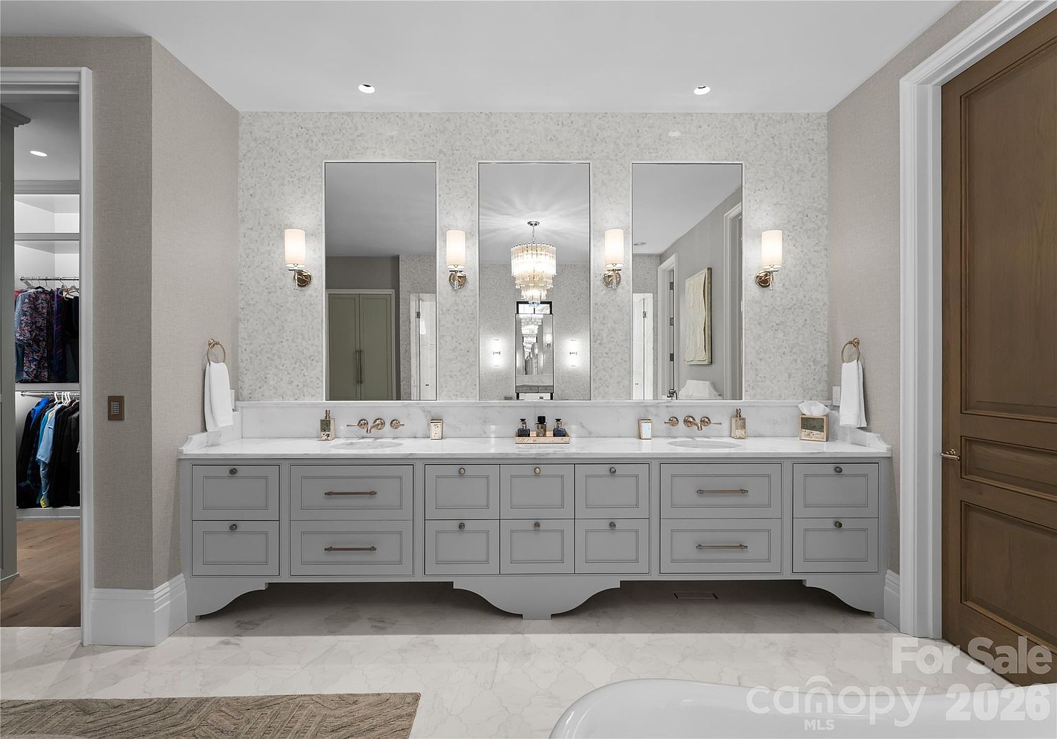 This primary bathroom features a long, double-vanity cabinet in a soft gray finish topped with a light-colored stone countertop. Three large, rectangular mirrors are mounted above the vanity, flanked by elegant wall-mounted light fixtures, while a central chandelier adds a touch of luxury. The space is finished with a light-toned, speckled wall treatment and includes a view into an adjacent walk-in closet on the left.