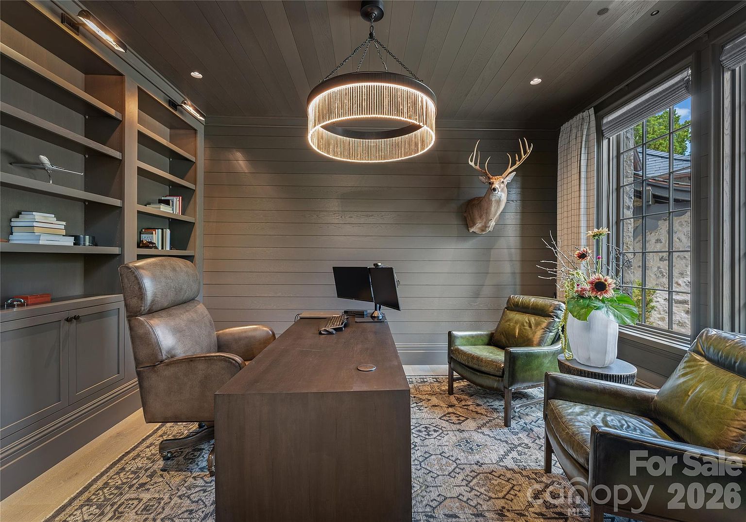 This sophisticated home office features dark, horizontal wood-paneled walls and a matching coffered ceiling, creating a moody and professional atmosphere. The room is anchored by a large, dark wood desk, a leather executive chair, and two green leather armchairs, all set upon a patterned area rug. A striking circular chandelier hangs above the desk, while built-in shelving and a mounted deer head add character to the space, which is illuminated by natural light from a window on the right.
