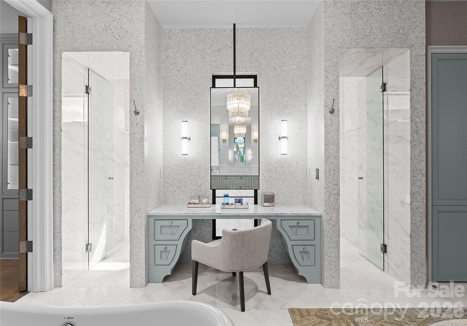 This elegant primary bathroom features a central vanity area with a light blue cabinet, a stone countertop, and a tall, framed mirror flanked by wall-mounted sconces. The space is framed by two glass-enclosed shower stalls with light-colored stone walls, creating a symmetrical and luxurious aesthetic. A light-colored upholstered chair sits at the vanity, and a portion of a white soaking tub is visible in the foreground, contributing to a spa-like atmosphere.