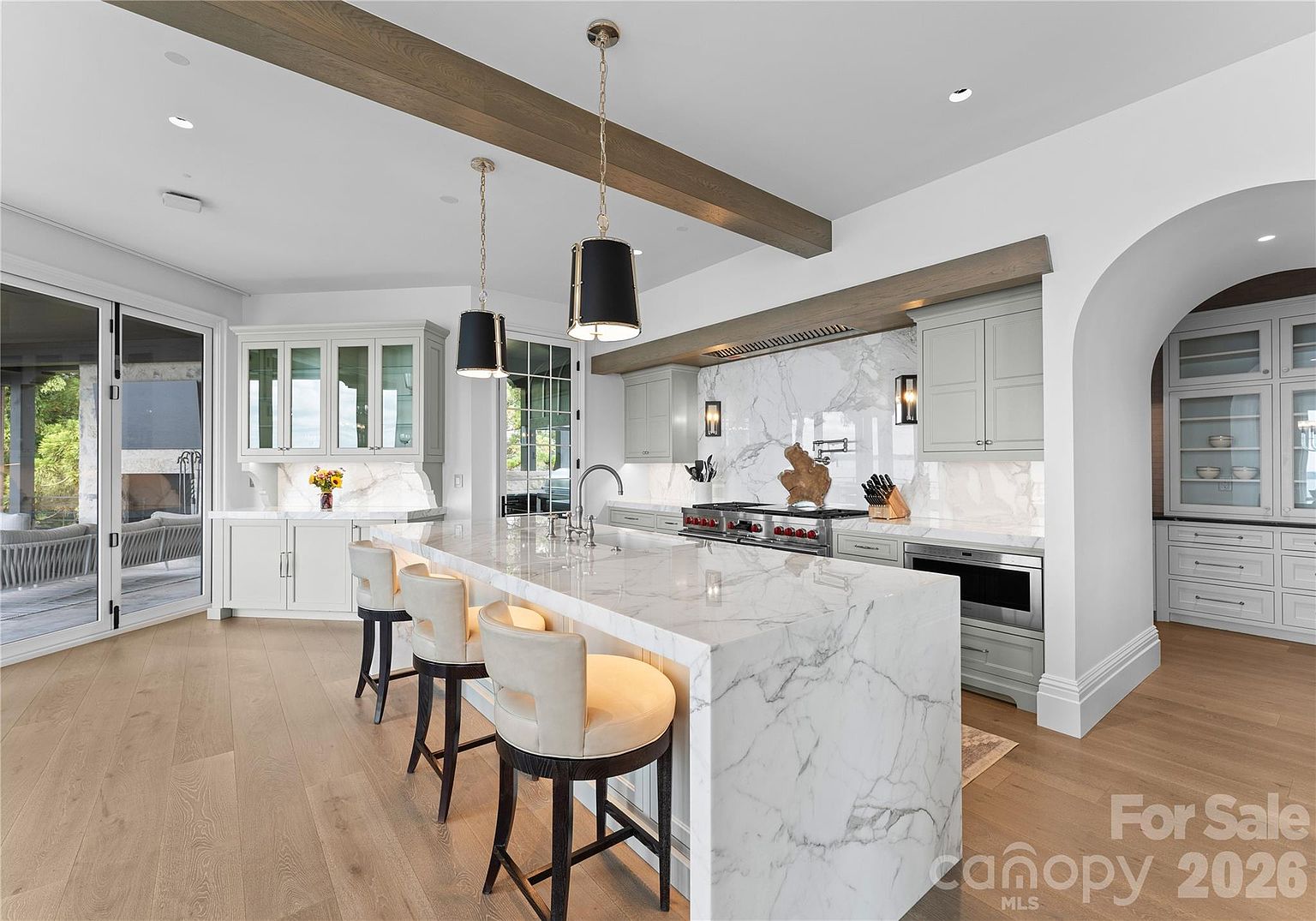 This bright and spacious kitchen features a large central island with a stone waterfall edge and three upholstered bar stools. The space is defined by light wood flooring, soft gray cabinetry, and a striking stone backsplash that extends behind the professional-grade range. Two black pendant lights hang above the island, while a wide glass door provides a view of the outdoor patio area.