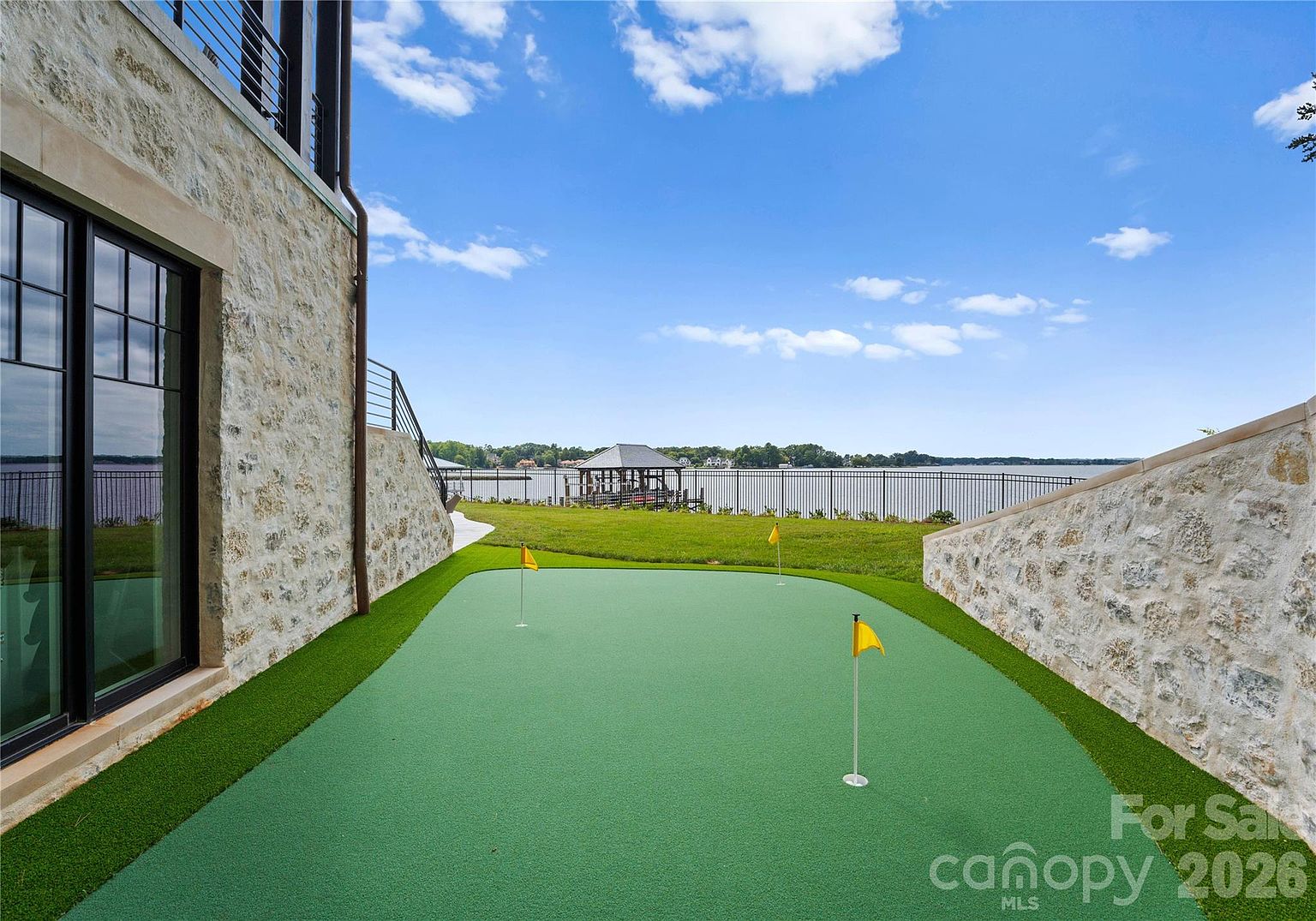 This outdoor space features a private putting green nestled between a stone-clad exterior wall and a retaining wall, creating a secluded recreational area. The green is equipped with multiple flags and overlooks a expansive lawn that leads to a black metal fence bordering a large lake. The perspective captures the transition from the home's architecture to the scenic waterfront, emphasizing a luxury lifestyle.