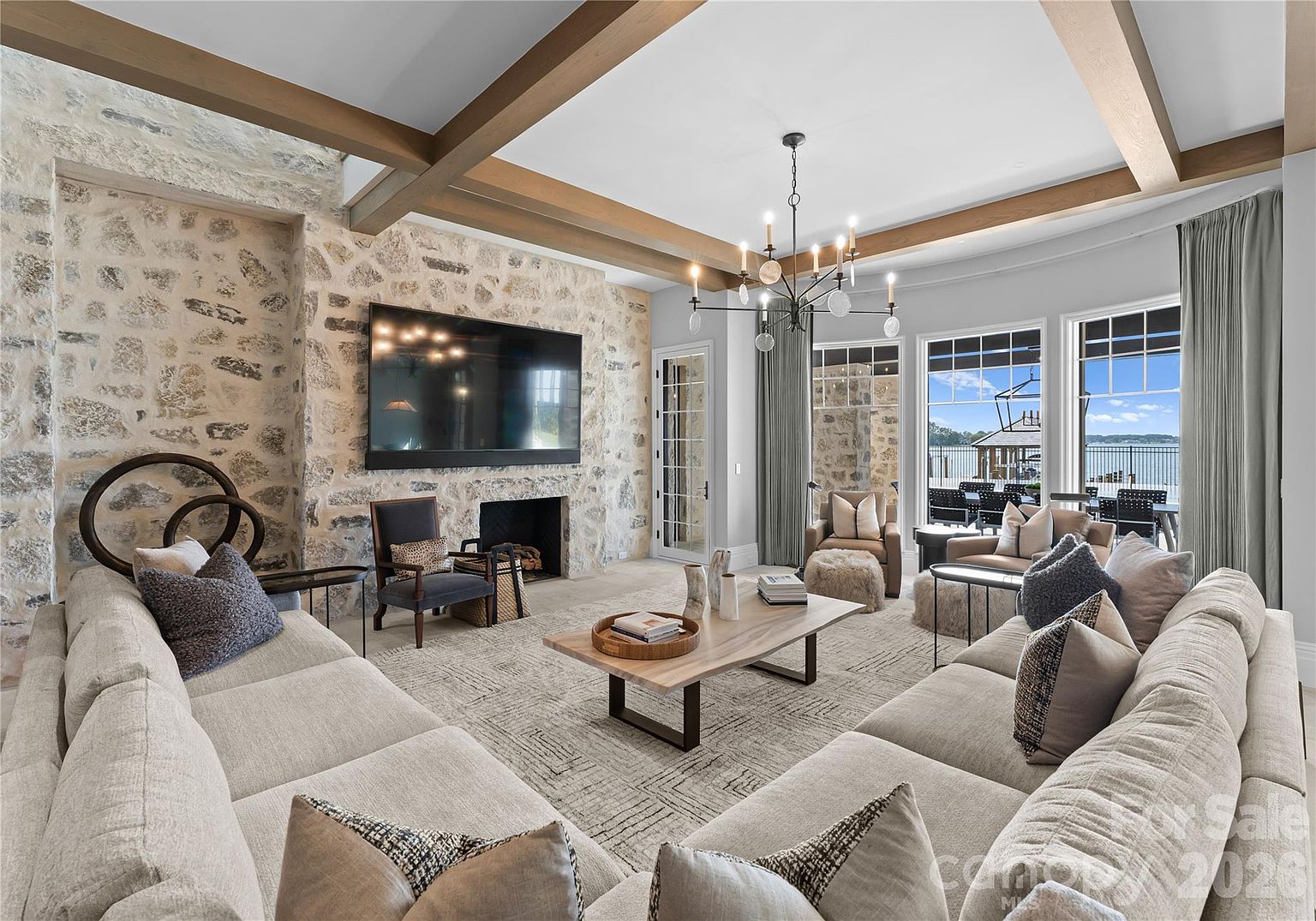 This spacious living room features a prominent stone-clad chimney breast that serves as the focal point, housing a television above the fireplace. The room is anchored by a large, light-colored sectional sofa and a central wooden coffee table, all set beneath exposed wooden ceiling beams. Large windows provide an expansive view of the water and outdoor patio, creating a bright and airy atmosphere with a sophisticated, rustic-modern aesthetic.