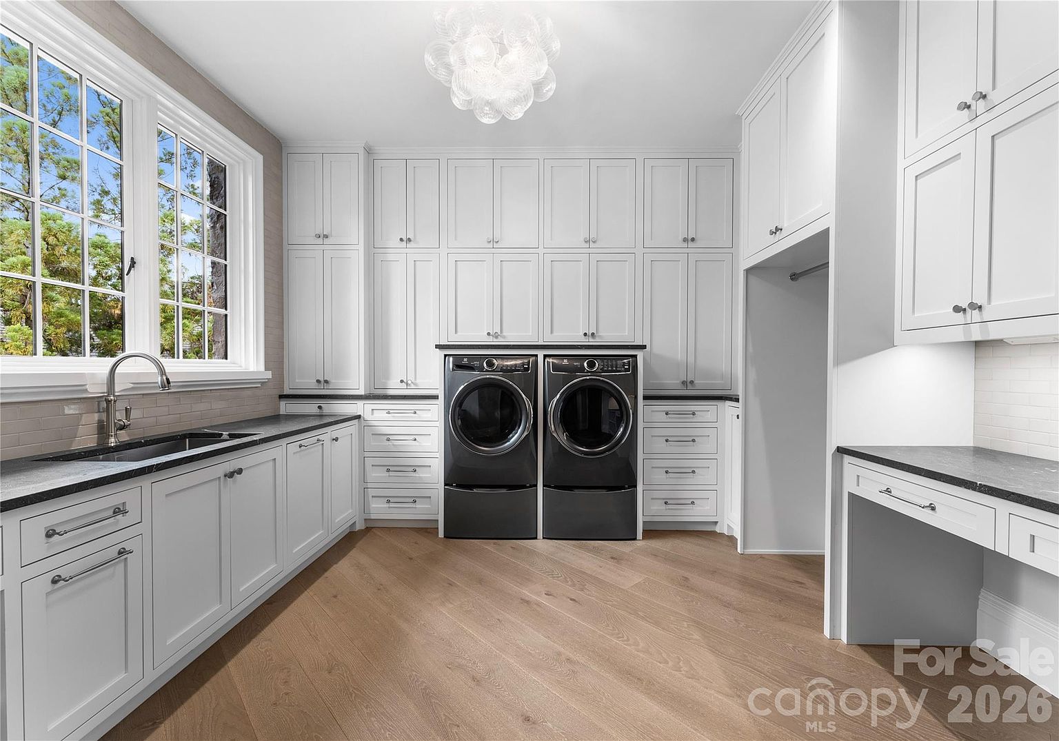 This bright and spacious laundry room features crisp white cabinetry, dark stone countertops, and light wood-toned flooring. The room is equipped with a deep sink, a pair of dark-toned laundry appliances, and a dedicated desk or folding area, all illuminated by a unique, multi-globe chandelier. The perspective is a wide-angle shot capturing the entire functional layout, emphasizing the clean, organized aesthetic of the space.