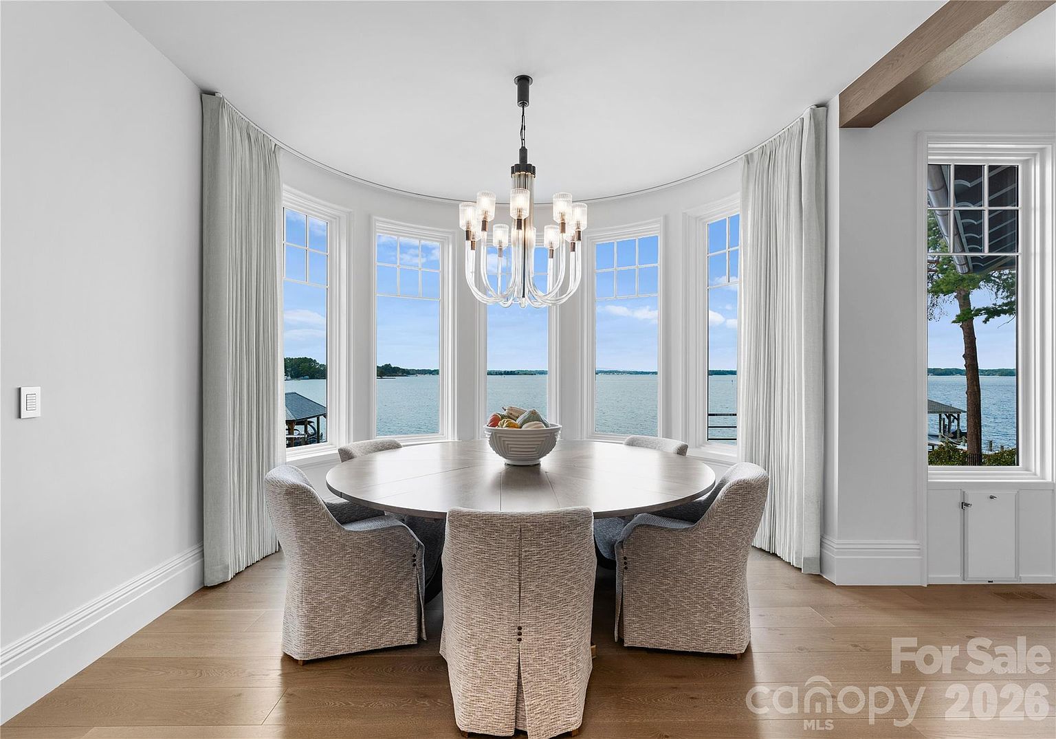This elegant dining room features a large, round wooden table surrounded by six upholstered chairs, centered beneath a modern multi-arm chandelier. The space is defined by a curved wall of large windows that provide expansive, unobstructed views of the water outside. Light wood flooring and neutral-toned walls create a bright, airy atmosphere, while floor-length curtains frame the scenic backdrop.