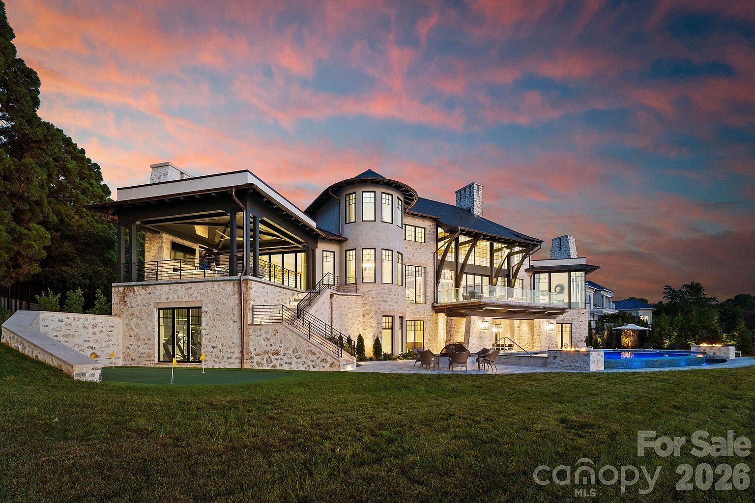 This expansive luxury estate features a sophisticated blend of stone and dark metal accents, showcasing a multi-level rear facade with a prominent circular tower element. The property includes a large covered patio, a tiered terrace with outdoor seating, and a custom-shaped swimming pool set against a vibrant sunset sky. A manicured lawn with a small putting green in the foreground completes this high-end outdoor living space.