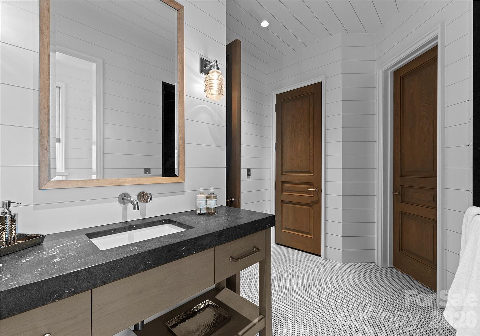 This modern bathroom features a floating vanity with a dark stone countertop and a rectangular undermount sink, paired with a large mirror framed in light wood. The walls are finished with horizontal white shiplap, and the floor is covered in small, white hexagonal tiles. Two dark wood doors lead to adjacent rooms, creating a clean, high-contrast aesthetic.