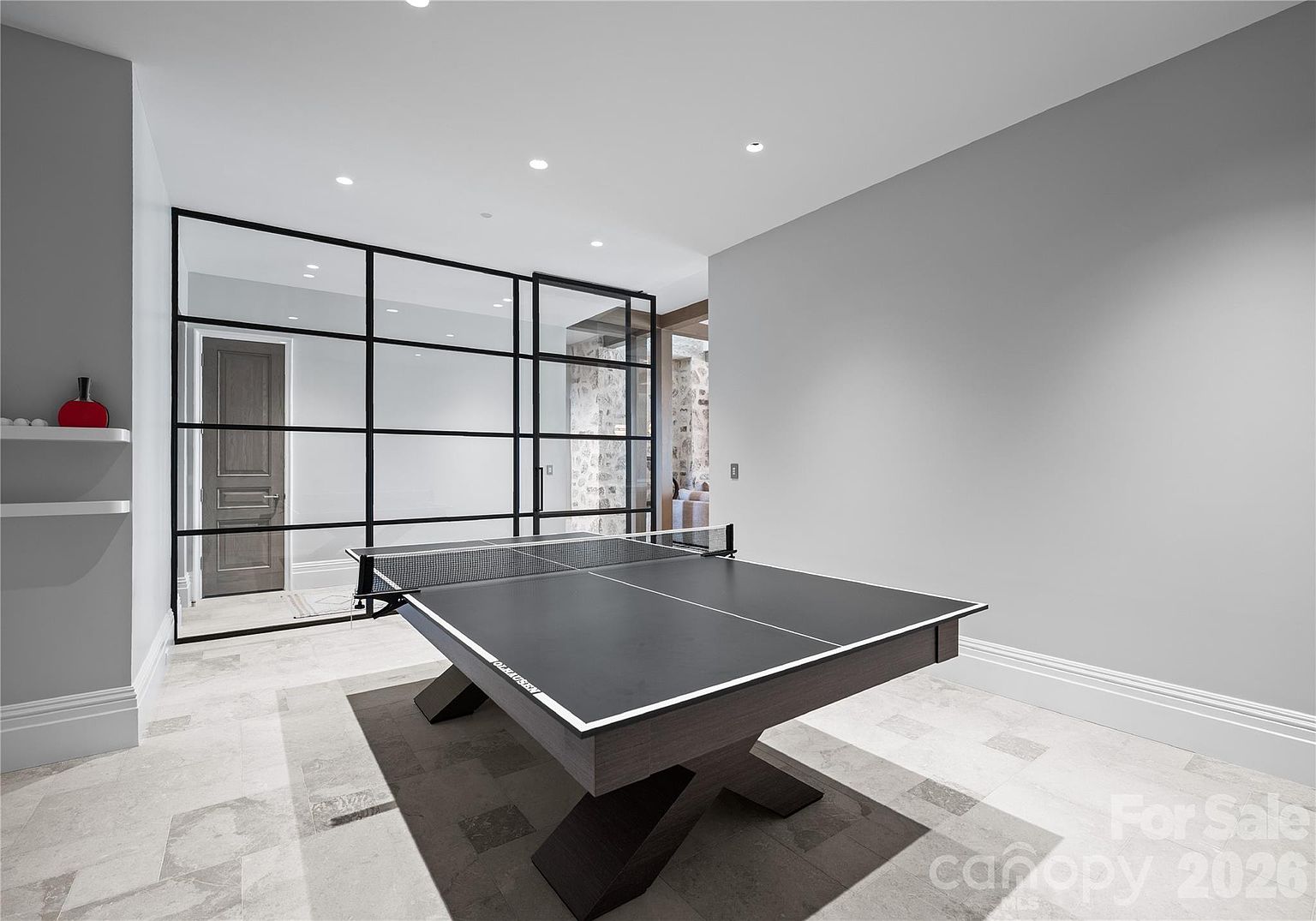 This modern recreation area features a sleek ping-pong table as the central focal point, set against a backdrop of light gray walls and recessed ceiling lighting. A striking black-framed glass partition separates the space from an adjacent room, adding a contemporary architectural element. The room is finished with large-format light-toned floor tiles, creating a clean and sophisticated atmosphere suitable for entertainment.