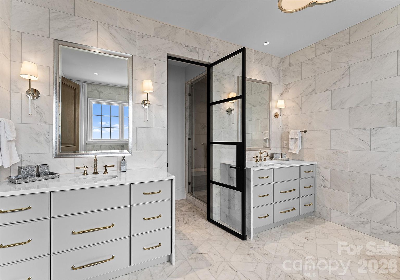 This luxurious primary bathroom features dual vanities with light-toned cabinetry, gold-toned hardware, and stone countertops. The space is finished with light-colored, veined stone wall tiles and matching patterned floor tiles, creating a cohesive and elegant aesthetic. A central black-framed glass door leads to a shower area, while wall-mounted sconces provide warm lighting on either side of the mirrors.