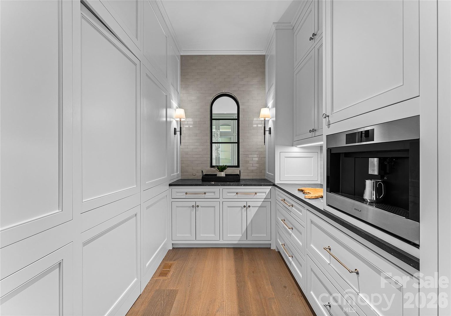 This elegant butler's pantry features crisp white cabinetry with gold-toned hardware and a dark stone countertop. A central arched window is framed by a light-colored tile backsplash and two wall-mounted sconces, creating a symmetrical and sophisticated focal point. The space is equipped with a built-in coffee machine, offering a seamless blend of luxury and functionality.