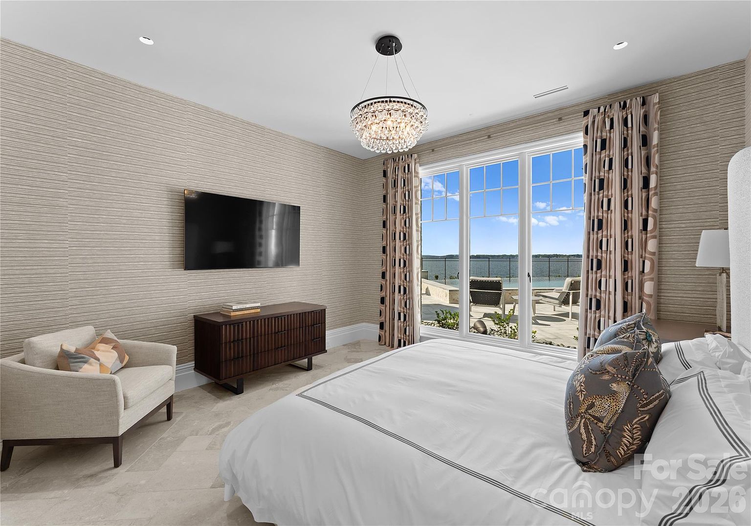 This elegant primary bedroom features a neutral color palette with textured wallpaper and a large bed adorned with patterned pillows. A sleek, dark wood dresser sits beneath a wall-mounted television, while a comfortable armchair provides a cozy seating area. Large glass doors offer a direct view of the outdoor patio and the lake beyond, filling the room with natural light.