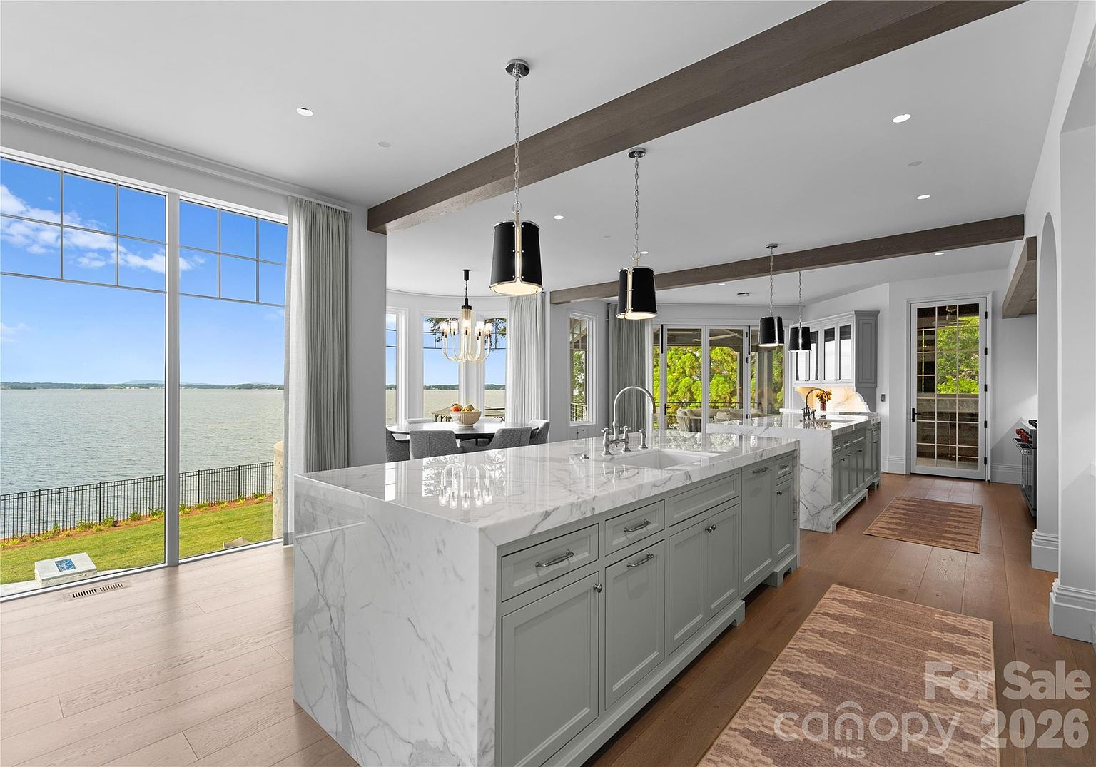This bright, open-concept kitchen features a large central island with a stone countertop and light-colored cabinetry, serving as the focal point of the space. Large windows provide expansive views of the lake, while dark wood ceiling beams add architectural interest to the room. The perspective is from the kitchen looking toward the dining area, emphasizing the seamless flow and airy, modern aesthetic of the home.