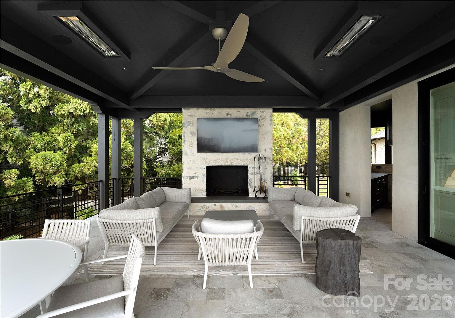 This covered outdoor living space features a dark-painted vaulted ceiling with a central ceiling fan and integrated heaters. A prominent stone-clad chimney breast serves as the focal point, housing a fireplace and a mounted television. The area is furnished with two light-colored sofas and a central chair, all arranged on a large area rug atop stone flooring, creating a sophisticated and inviting atmosphere for relaxation.