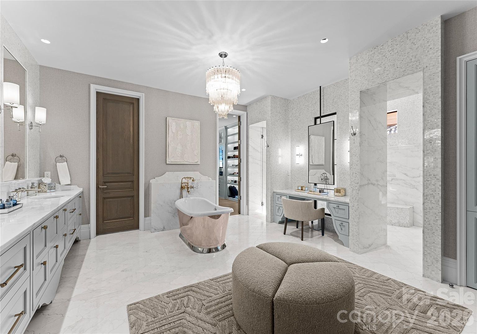This luxurious primary bathroom features a freestanding metallic-finished tub set against a stone-clad wall, complemented by a double vanity with ample storage and a dedicated makeup vanity area. The space is illuminated by a grand crystal chandelier and elegant wall sconces, while a large walk-in shower is visible through a wide stone-trimmed opening. The room is finished with light-toned stone flooring and a neutral color palette, creating a sophisticated and serene atmosphere.