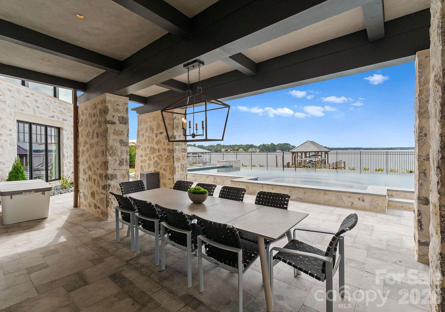 This covered outdoor dining area features a large rectangular table surrounded by eight modern chairs, set beneath a dark-beamed ceiling with a prominent geometric pendant light. The space is framed by rustic stone pillars and opens directly onto a sleek infinity-edge pool overlooking a calm lake. The perspective captures a seamless transition from the shaded patio to the expansive waterfront view, highlighting a luxurious and tranquil outdoor living environment.