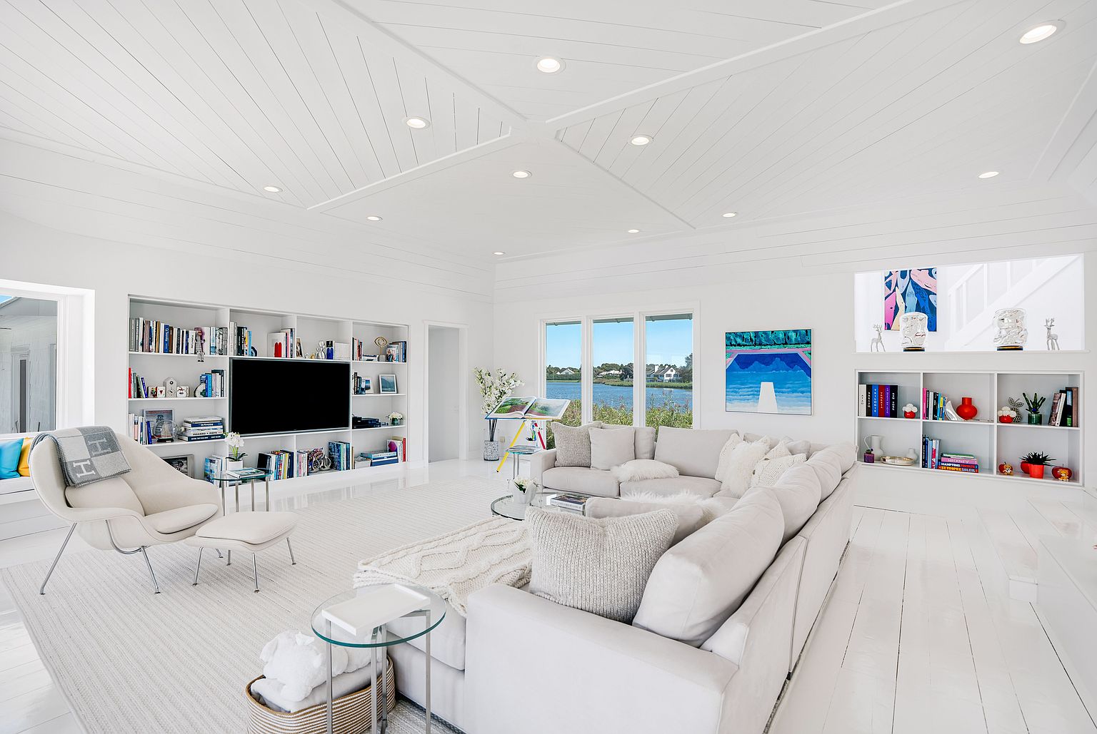 This bright and airy living room features a crisp, all-white aesthetic with a large sectional sofa and a modern lounge chair with an ottoman. The space is anchored by built-in shelving units flanking a television and a decorative display area, while large windows provide a scenic view of the water outside. The room is finished with a unique geometric ceiling design and recessed lighting, creating a clean, sophisticated, and inviting atmosphere.