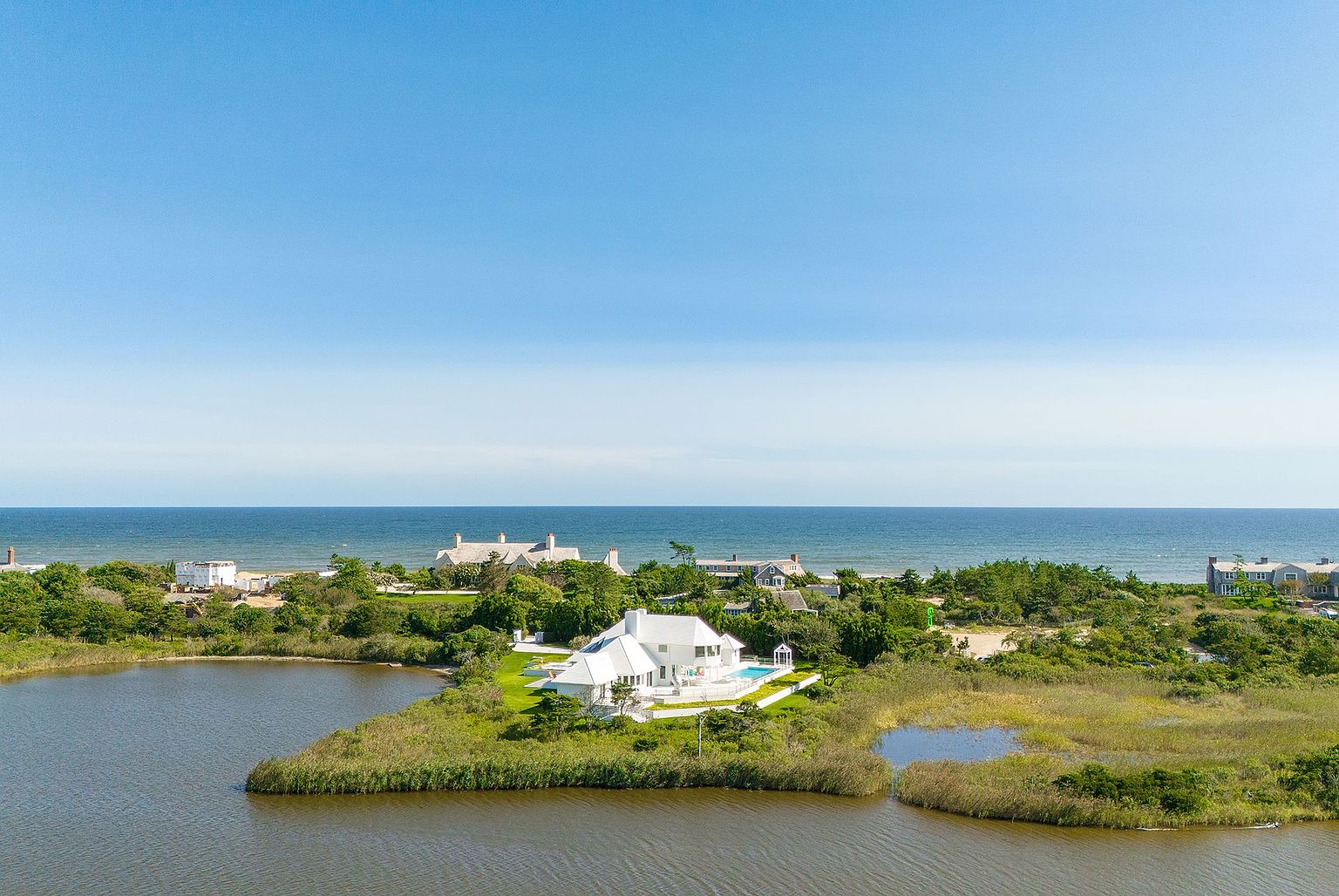 This high-angle aerial perspective captures a pristine white coastal residence situated on a lush, grassy peninsula surrounded by tranquil marshland and water. The property features a private swimming pool, manicured lawns, and a clean, modern architectural aesthetic that contrasts beautifully with the natural landscape. In the background, the expansive blue ocean meets the horizon, emphasizing the home's secluded and serene waterfront location.