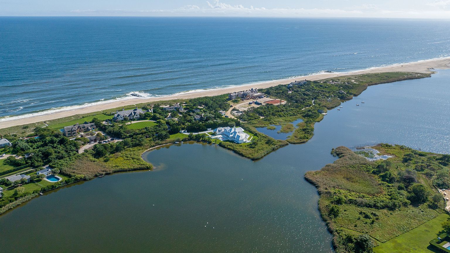 This high-altitude aerial perspective captures a luxurious coastal estate situated on a narrow strip of land between the ocean and a tranquil inlet. The property features multiple sprawling residences with manicured lawns, private pools, and lush landscaping, all set against the backdrop of the vast blue sea. The composition highlights the exclusive, secluded nature of the waterfront location, emphasizing the seamless integration of high-end architecture with the natural coastal environment.
