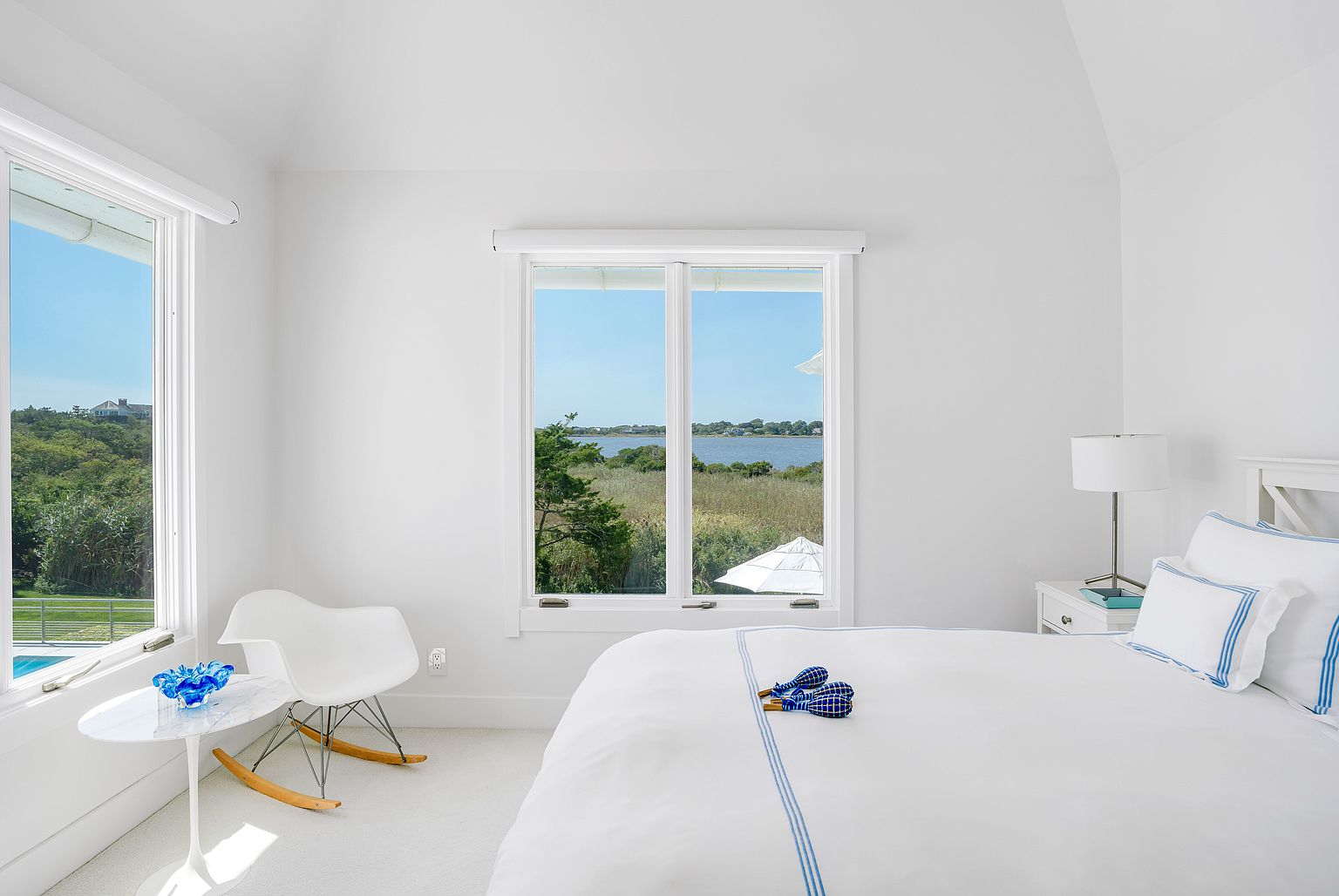 This bright and airy bedroom features a crisp, coastal-inspired aesthetic with white walls and plush carpeting. A comfortable bed with white linens and blue-striped accents sits beside a bedside table with a lamp, while a modern rocking chair and small side table are positioned near large windows that frame a scenic water view. The room is filled with natural light, creating a serene and inviting atmosphere.