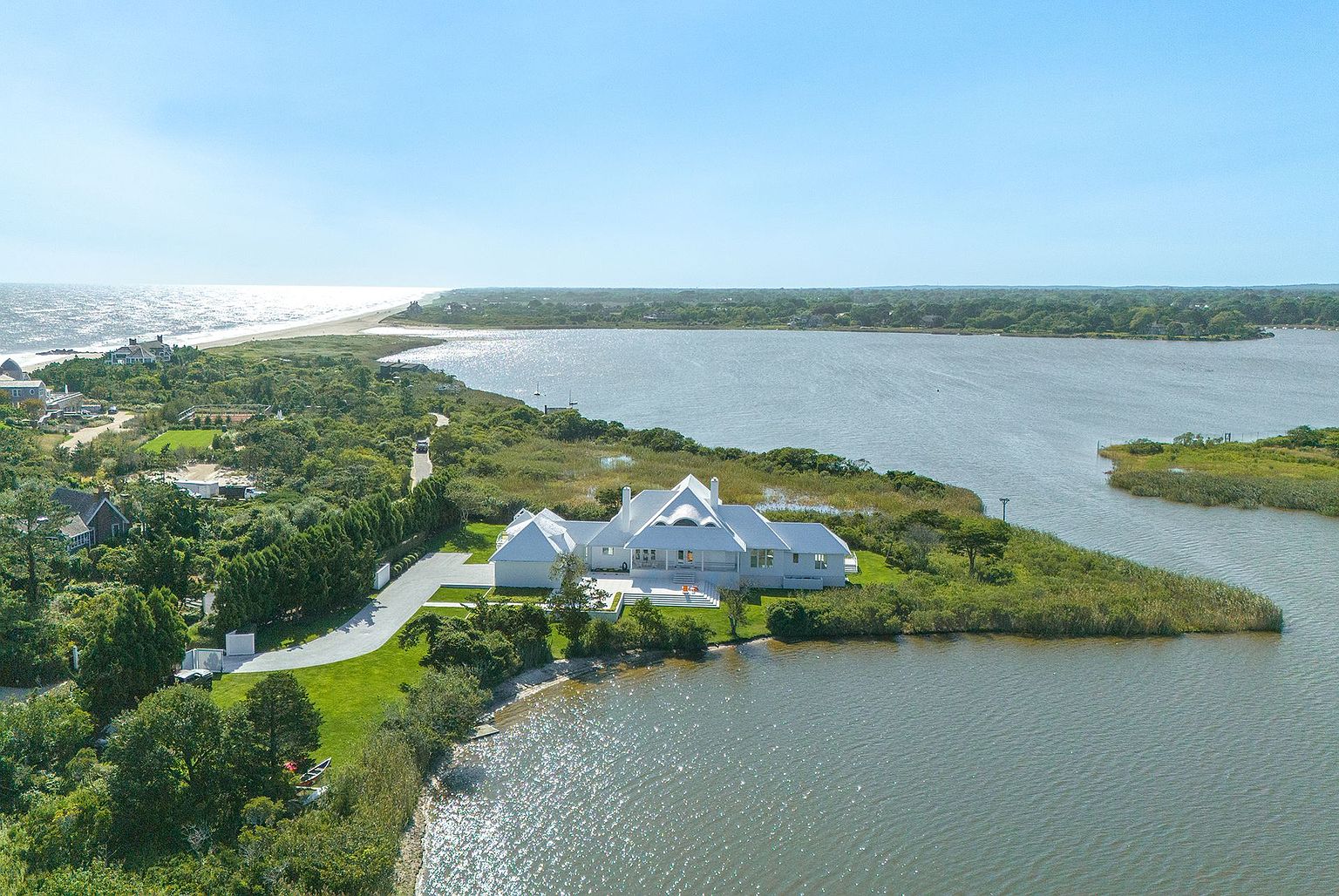 This high-angle aerial view captures a sprawling white coastal residence situated on a secluded peninsula surrounded by water. The property features a manicured lawn, a long private driveway, and a prominent white roofline, set against a backdrop of lush greenery and the distant ocean coastline. The perspective emphasizes the home's expansive waterfront location and its serene, private setting.