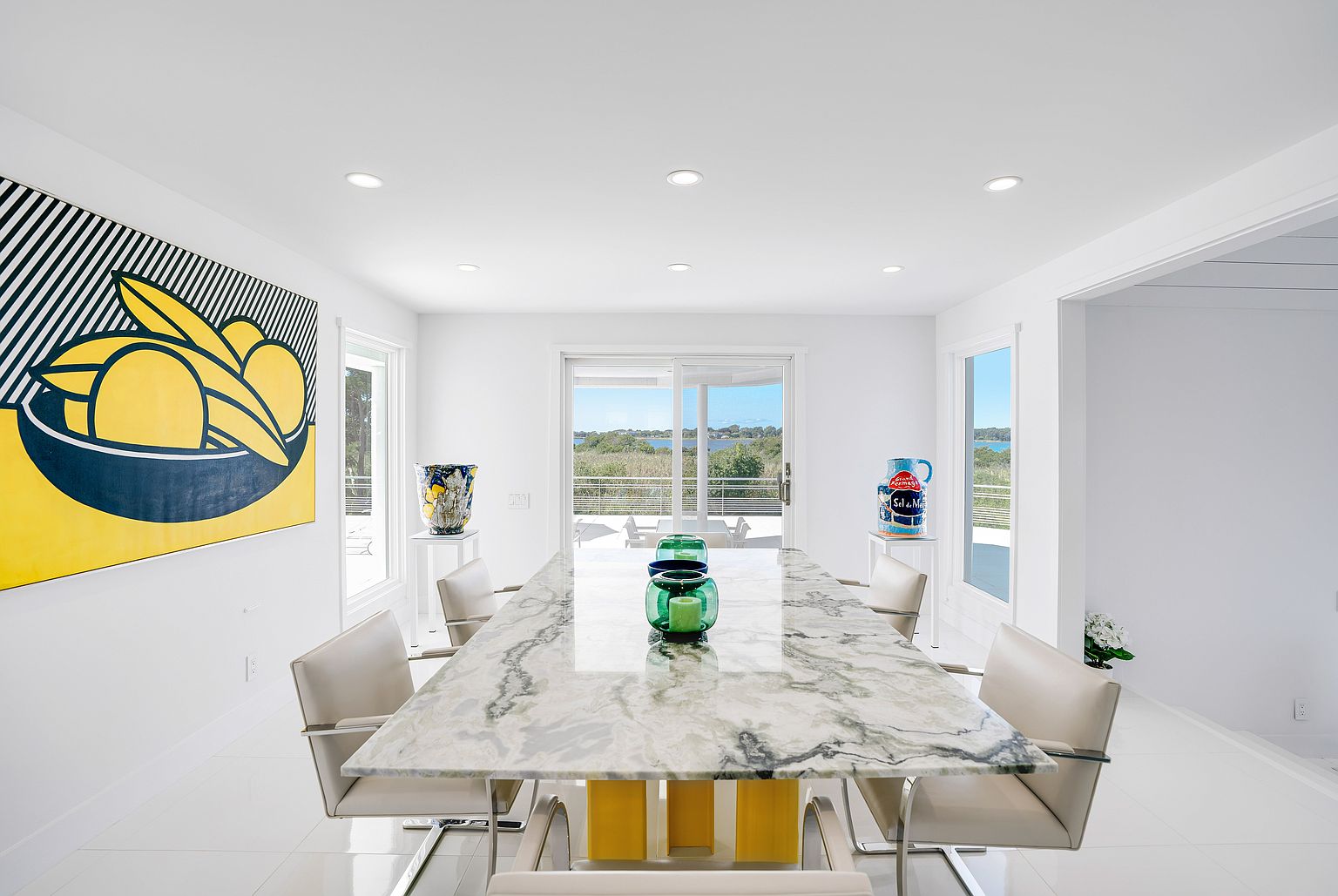 This bright and airy dining room features a large stone-topped table with a bold yellow base, surrounded by modern, neutral-toned chairs. A striking, large-scale graphic art piece depicting stylized fruit hangs on the left wall, while decorative ceramic vessels sit on pedestals near the windows. The room is bathed in natural light from large sliding glass doors that provide a clear view of the outdoor landscape and water beyond.