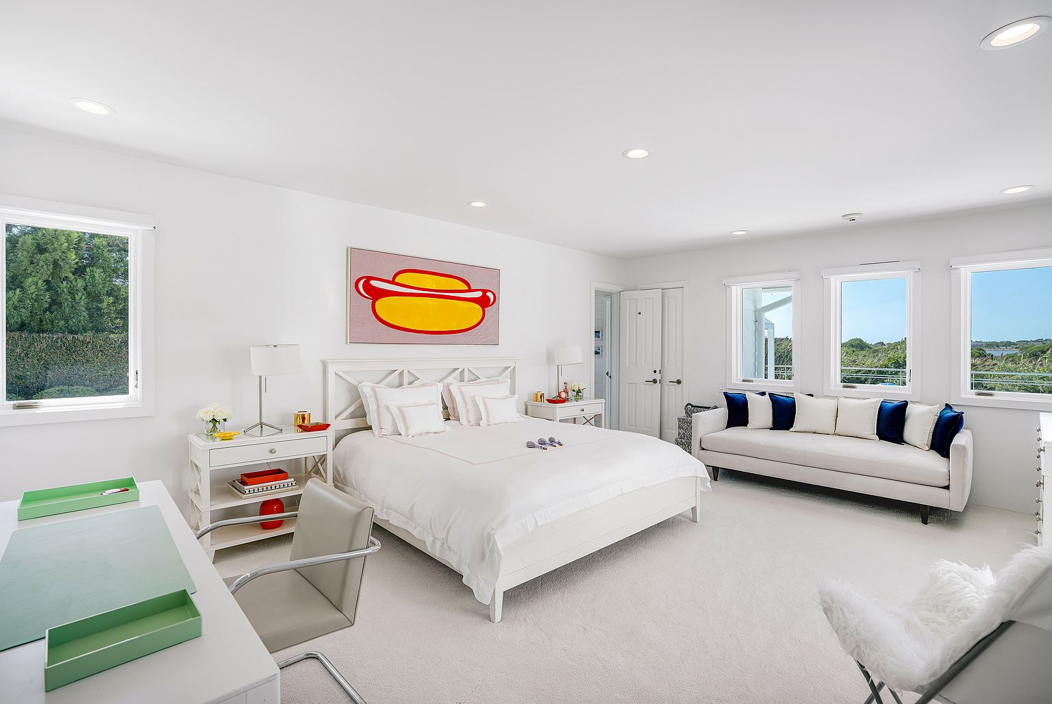 This bright and airy primary bedroom features a crisp white color palette, a central bed with a decorative hot dog art piece above it, and a comfortable sofa positioned near large windows that offer scenic outdoor views. A dedicated workspace with a desk and chair is situated in the foreground, while the room is illuminated by recessed lighting and natural light. The overall aesthetic is clean, modern, and spacious, creating a serene retreat.