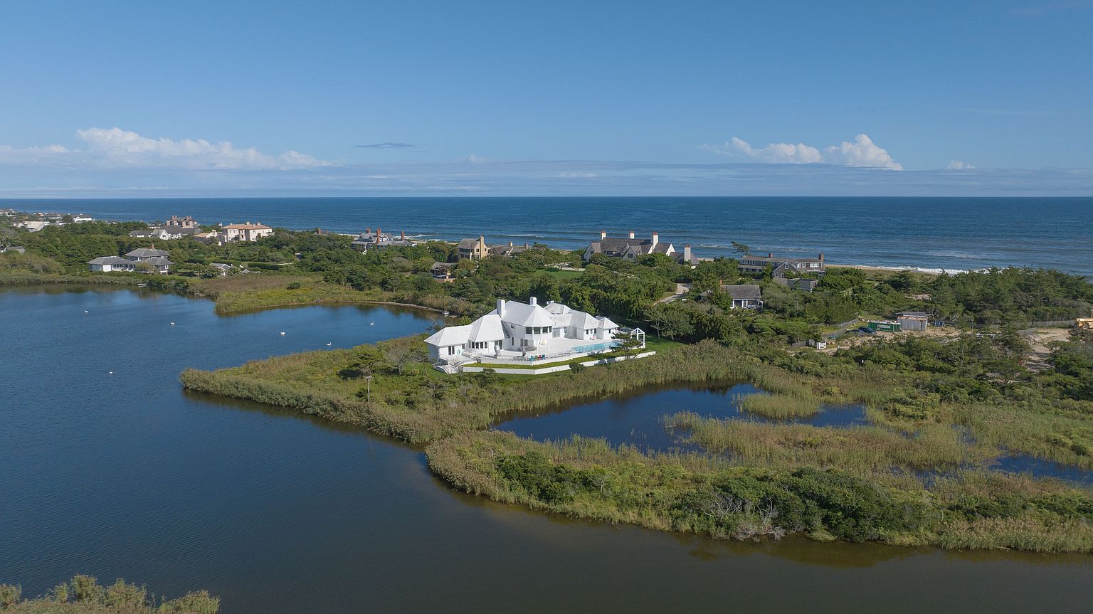 This high-angle aerial view captures a sprawling white coastal estate situated on a secluded peninsula surrounded by marshland and water. The property features a multi-gabled roofline, a private swimming pool, and expansive outdoor living areas that overlook the nearby ocean coastline. The surrounding landscape is lush with native greenery, emphasizing the home's private and serene waterfront location.