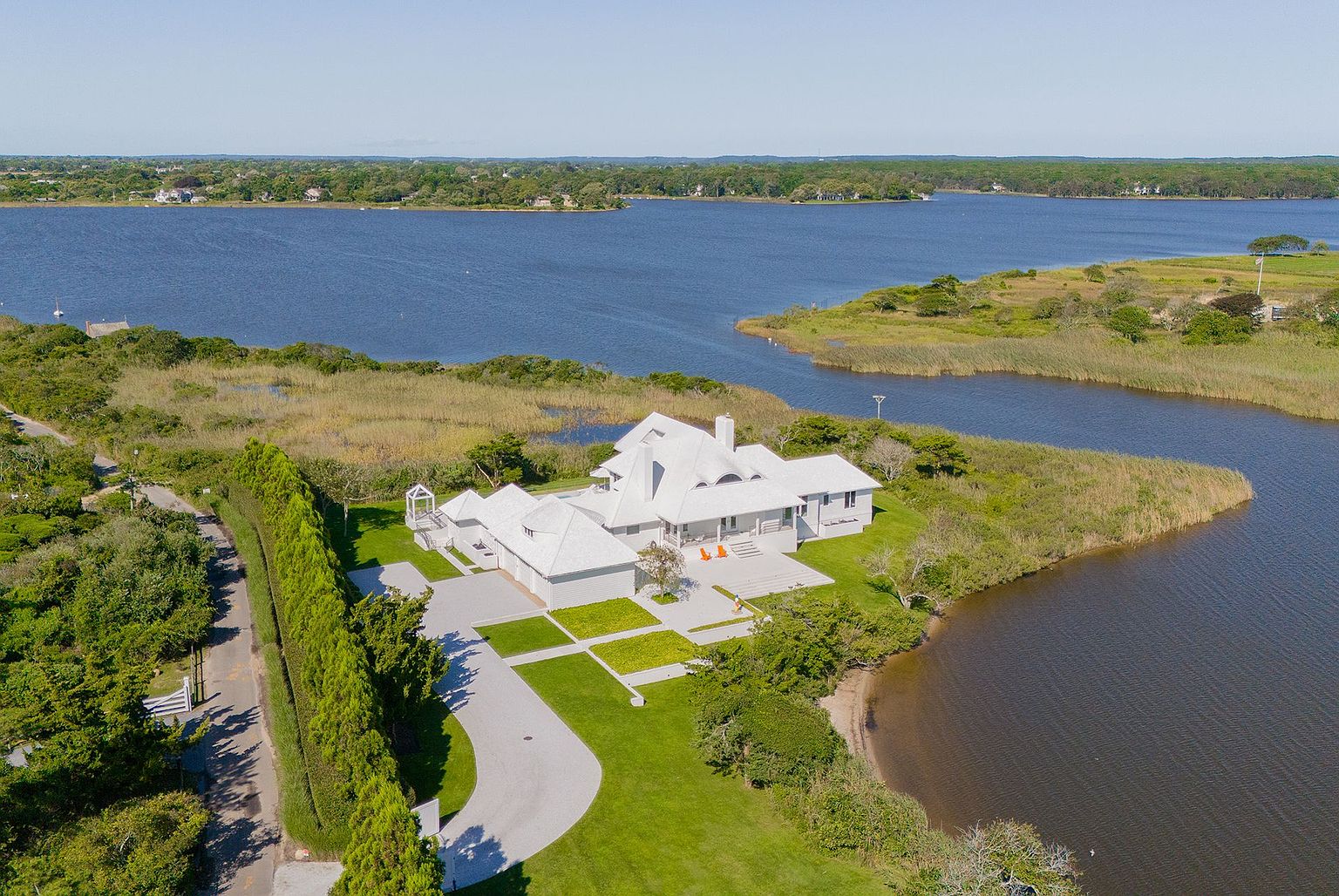 This high-angle aerial shot captures a sprawling, light-colored coastal residence situated on a point of land surrounded by water and marshland. The property features a multi-gabled roofline, a paved driveway leading to a garage, and a spacious rear patio area overlooking the water. The lush green lawn and natural landscape create a serene, secluded atmosphere, highlighting the home's prime waterfront location.