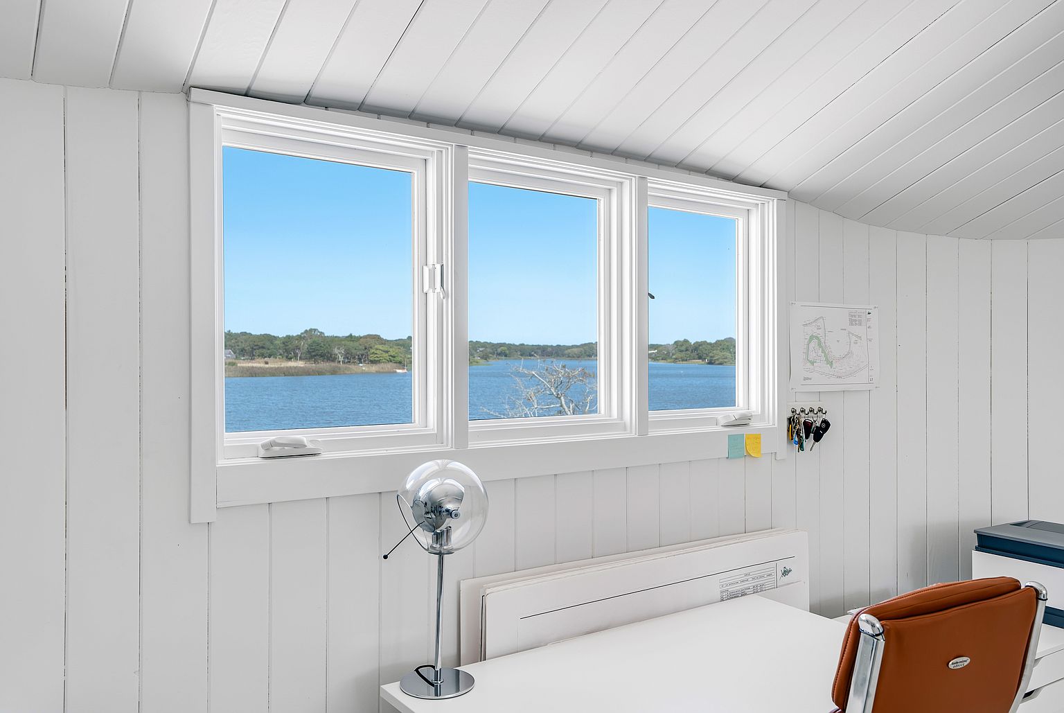 This bright home office features white vertical wood-paneled walls and a large triple-window configuration that frames a serene waterfront view. A minimalist white desk is positioned beneath the windows, accompanied by a modern brown leather chair and a sleek, spherical desk lamp. The space offers a clean, tranquil atmosphere perfect for focused work, with a small map and keys hanging on the wall adding a personal touch.
