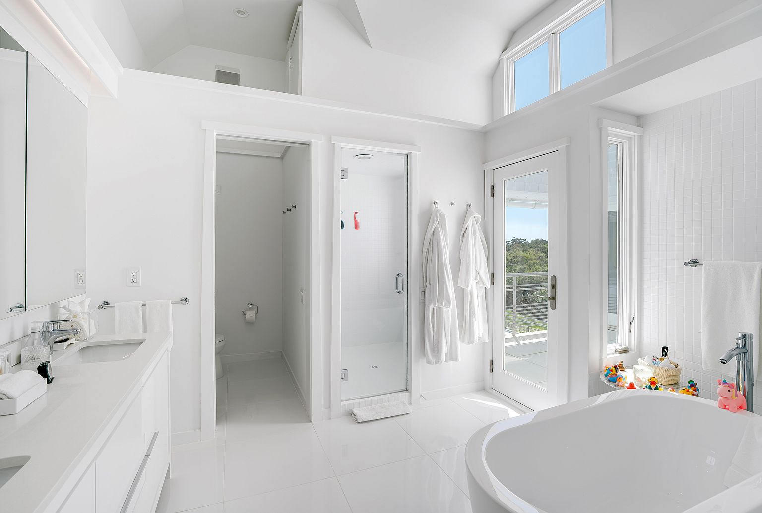 This bright, modern primary bathroom features a crisp white aesthetic with a double vanity, a separate enclosed toilet area, and a glass-walled shower. A deep soaking tub sits in the foreground, while a glass door provides direct access to an outdoor balcony, allowing for abundant natural light. The space is clean and minimalist, accented by white robes hanging near the shower and a small collection of bath toys near the tub.