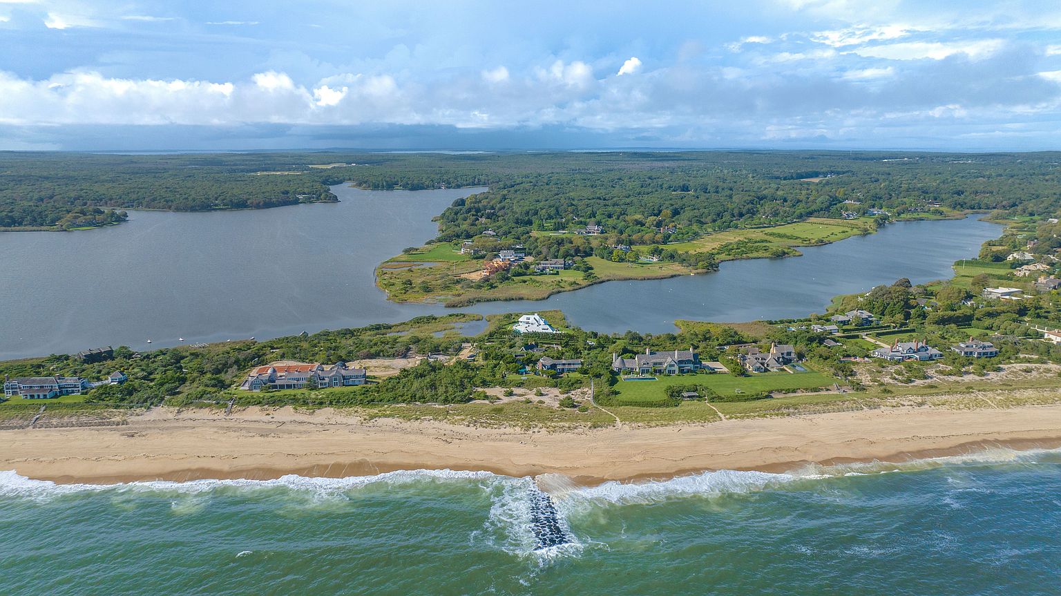 This expansive aerial view captures a prime coastal property situated on a narrow strip of land between the ocean and a large inland bay. The scene highlights several luxury waterfront estates with manicured lawns, private beach access, and a stone jetty extending into the surf. The composition emphasizes the unique geography and the serene, high-end lifestyle offered by this exclusive shoreline location.