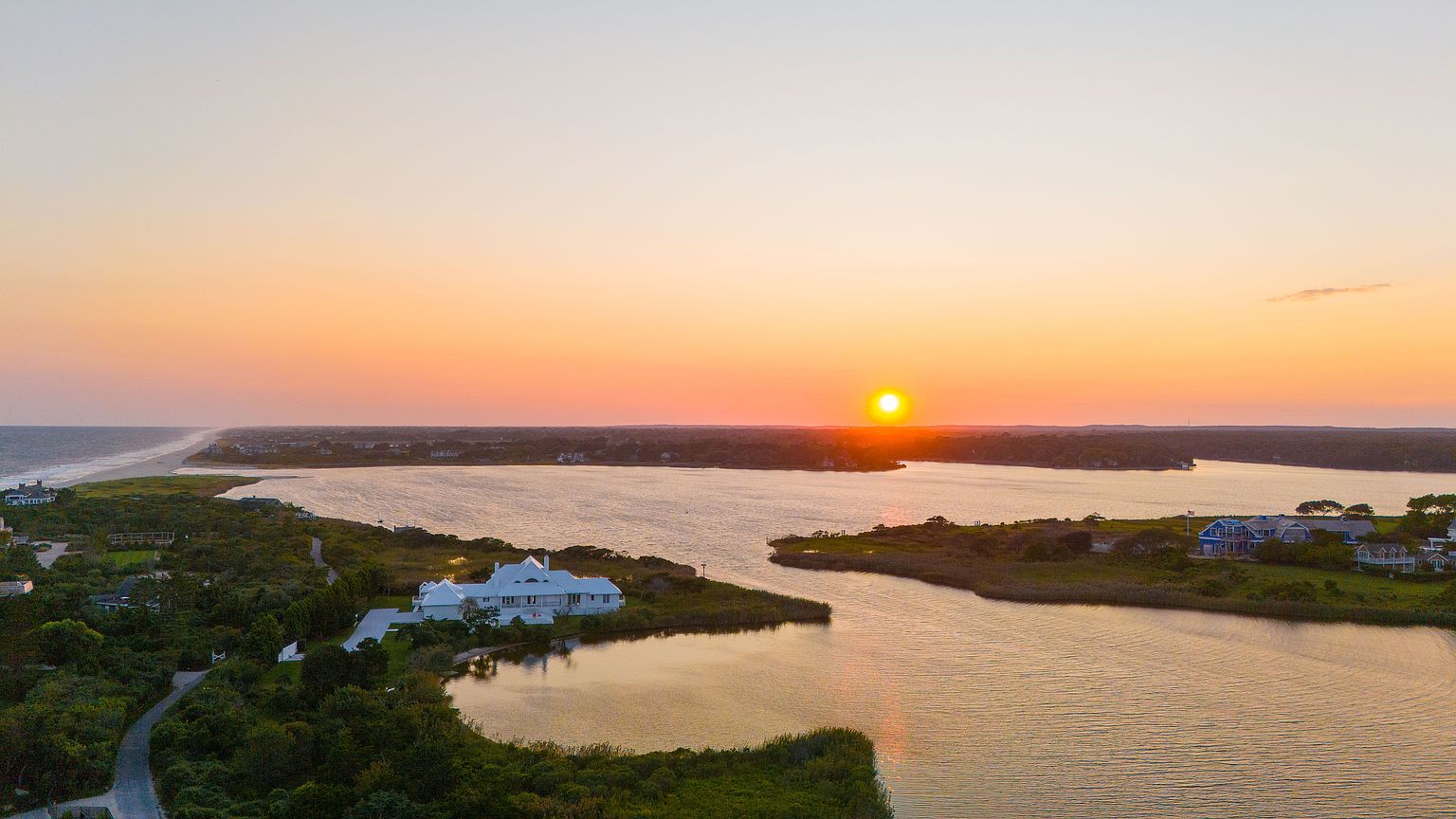 This stunning aerial perspective captures a sprawling coastal estate at sunset, overlooking a serene inlet and the distant ocean horizon. The property features a prominent white residence nestled amidst lush greenery, with expansive water views that create a tranquil and luxurious atmosphere. The golden hour light bathes the entire landscape in warm hues, highlighting the unique waterfront location and the peaceful, secluded setting of the home.