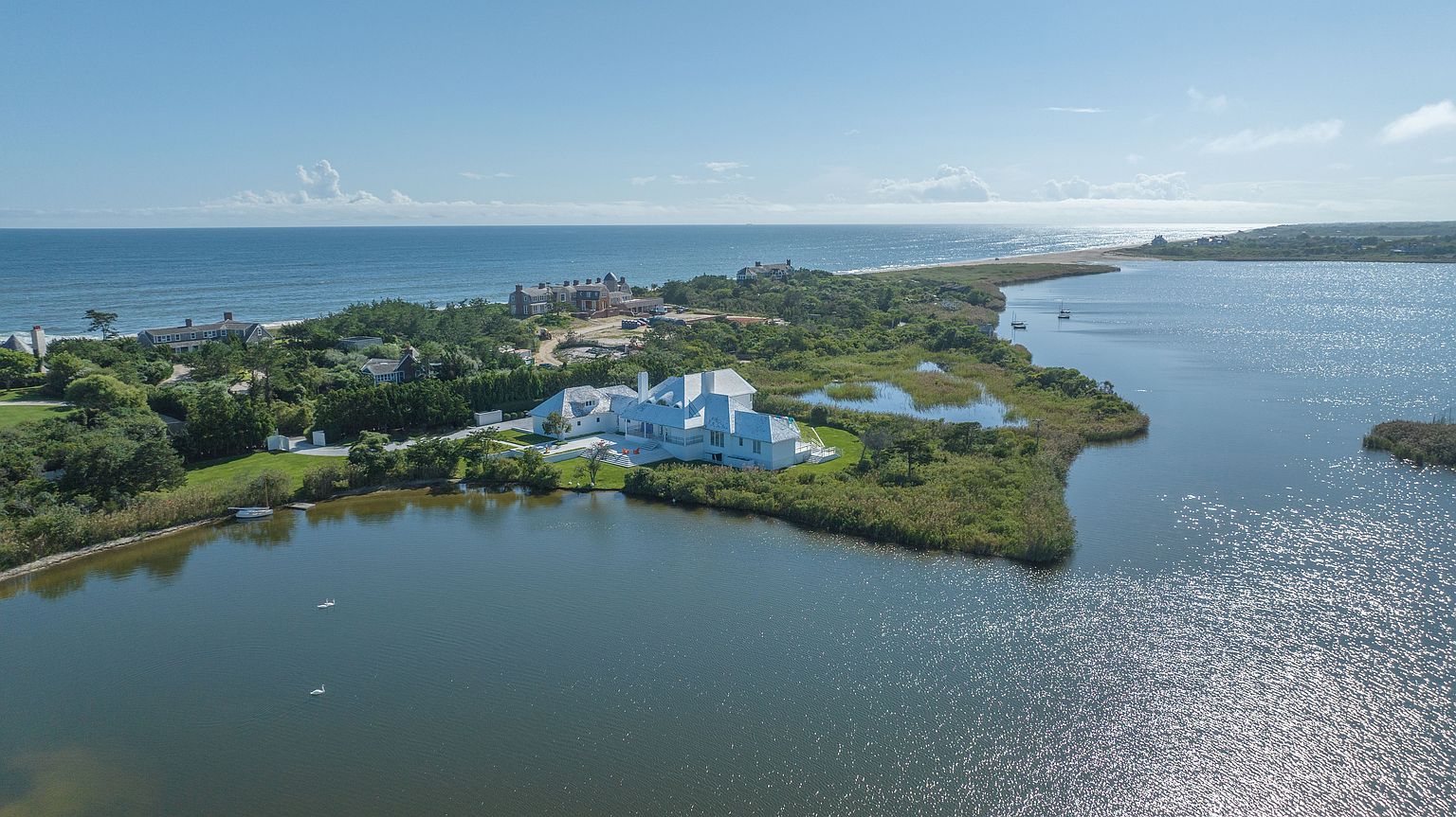This high-angle aerial view captures a sprawling, modern white estate situated on a lush, green peninsula surrounded by a serene body of water. The property features a multi-gabled roofline and expansive grounds that blend seamlessly into the natural coastal landscape, with the open ocean visible in the distance. The perspective emphasizes the secluded, waterfront location and the architectural harmony between the residence and its tranquil environment.