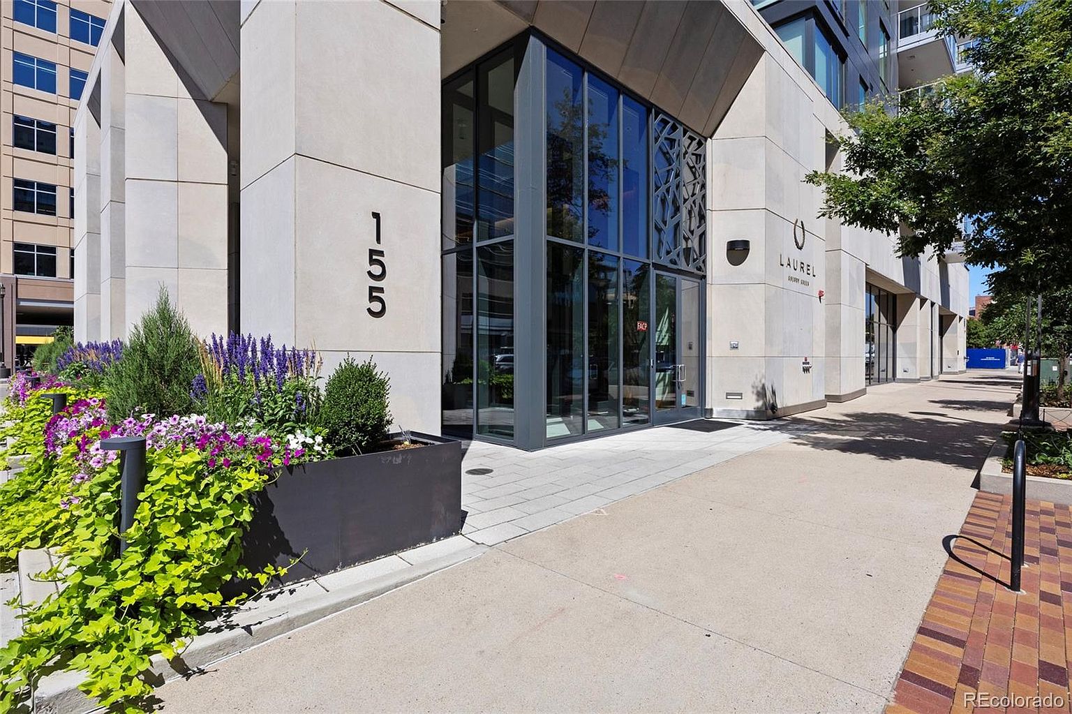 The image showcases the exterior entryway of a modern building, featuring a glass-enclosed entrance with a geometric design and the building's address prominently displayed as '155'. A well-maintained flower bed adds a touch of nature to the urban setting, and the building's facade is constructed with light-colored stone, creating a clean and sophisticated aesthetic. The perspective is at street level, emphasizing the building's accessibility and inviting atmosphere.