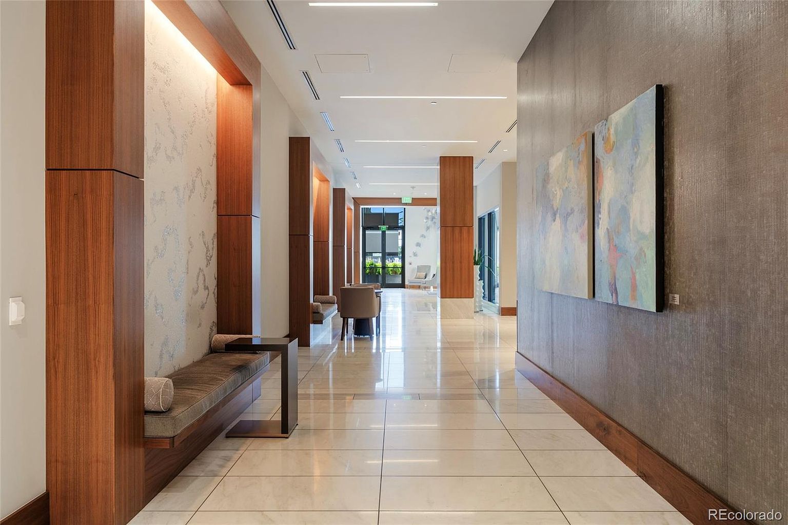 This is an interior shot of a modern hallway featuring marble tile flooring, wood accents, and neutral-toned walls. The hallway is well-lit with recessed lighting and natural light coming from the end of the hall. There are benches along the walls, and artwork is displayed on the right wall, creating a sophisticated and inviting atmosphere.