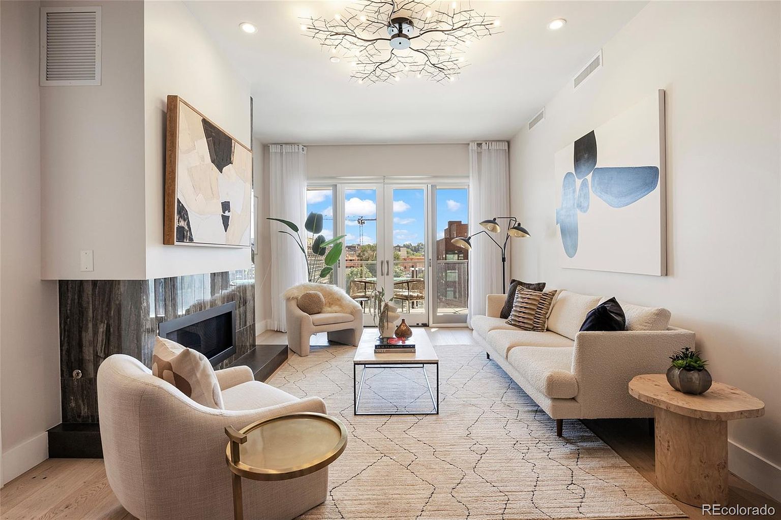 This is a bright and airy living room featuring a modern aesthetic with neutral tones and stylish furnishings. The room includes a fireplace, comfortable seating, contemporary artwork, and access to an outdoor balcony through glass doors, creating an inviting and sophisticated space. A unique, branch-like chandelier adds a touch of artistic flair to the room.