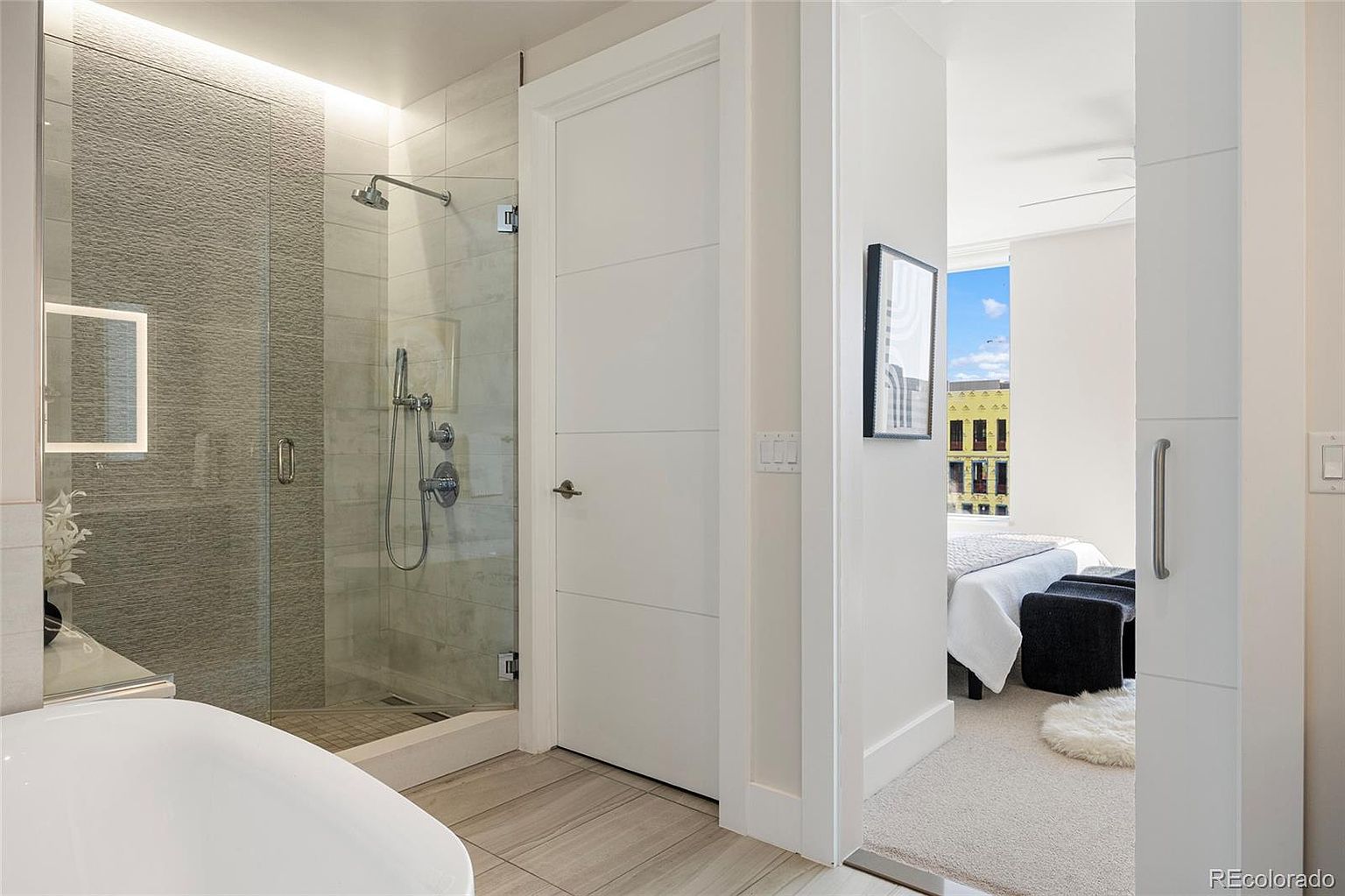 This is a bright and modern primary bathroom featuring a glass-enclosed shower with textured gray tile and a rainfall showerhead. The bathroom has light wood-look tile flooring and a white door with horizontal paneling. The view extends into a bedroom with a bed and seating area, creating a sense of spaciousness and connectivity.
