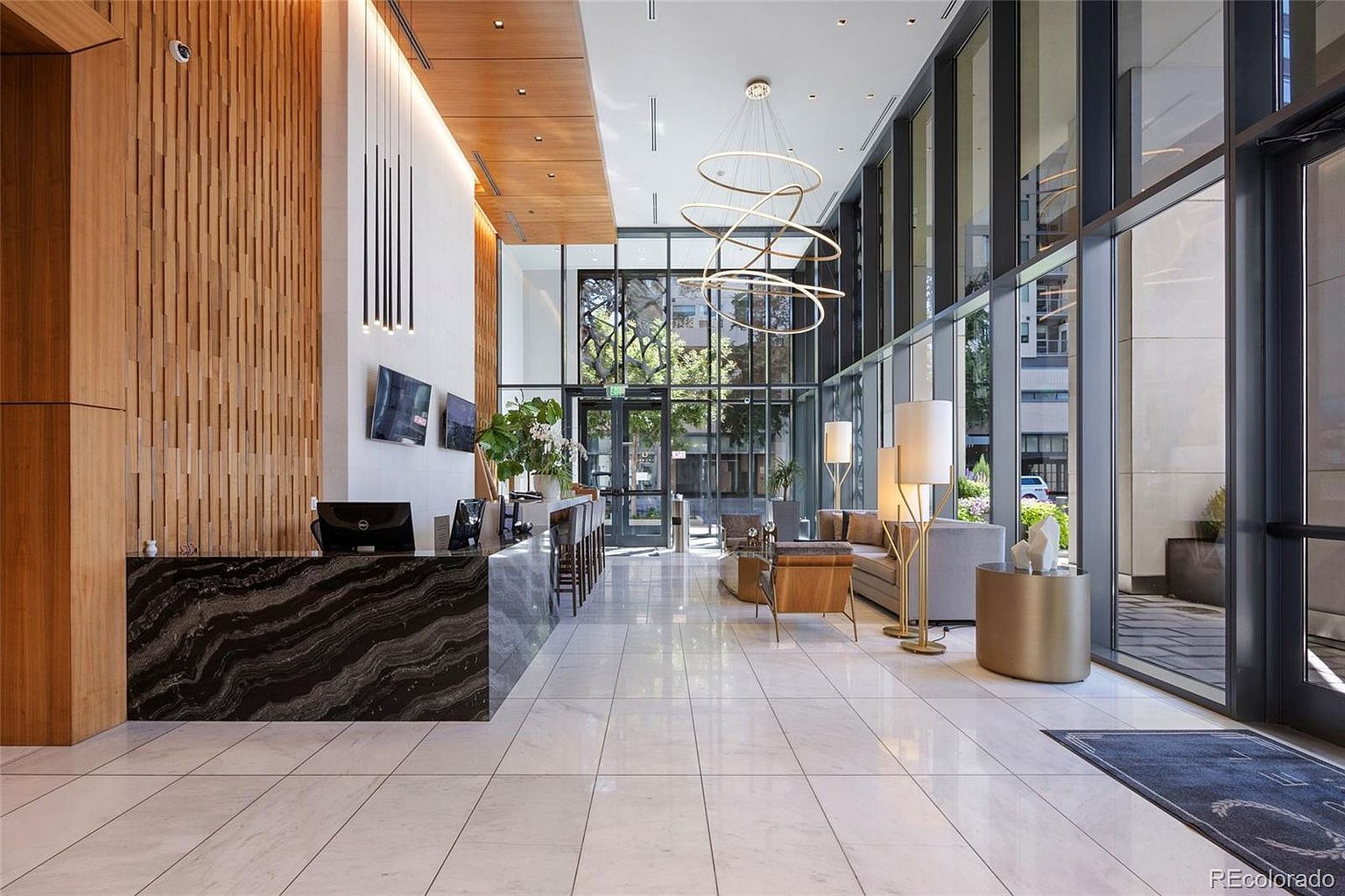 This is an interior shot of a modern building lobby. The space features a reception desk with a dark marble countertop, a wooden slat wall, and a seating area with contemporary furniture. Large windows provide ample natural light, and a unique chandelier hangs from the ceiling, creating a luxurious and welcoming atmosphere.