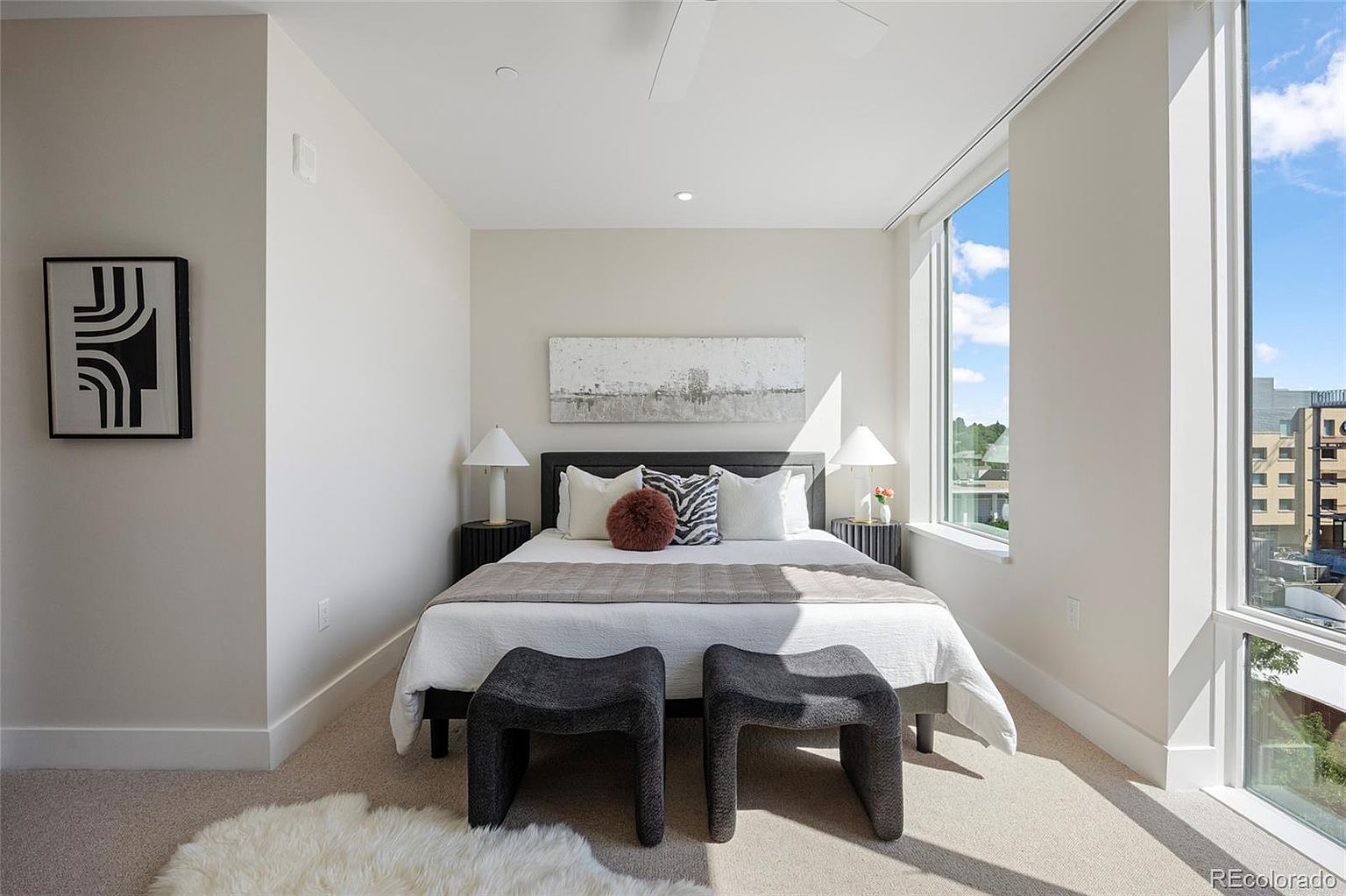 This is a well-lit primary bedroom featuring a bed with white linens, decorative pillows, and a gray throw. Two dark-colored stools are placed at the foot of the bed. The room has a modern aesthetic with neutral wall colors, a large window providing natural light, and artwork on the walls, creating a serene and inviting atmosphere.