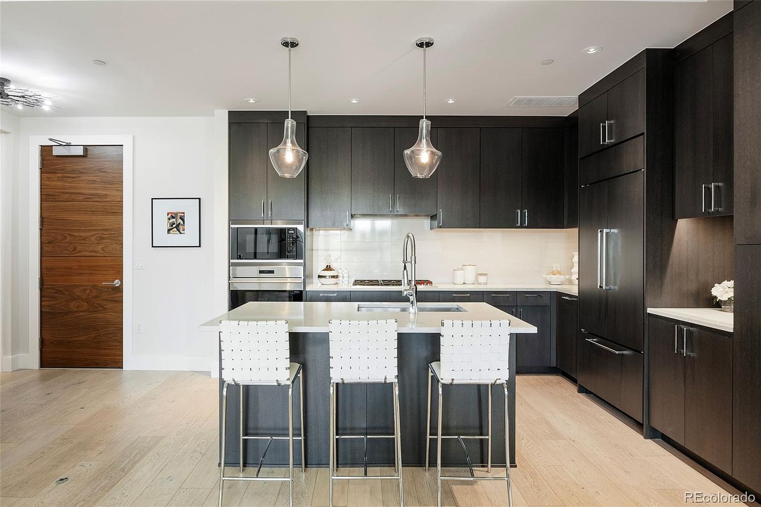 This is a modern kitchen featuring dark wood cabinetry, stainless steel appliances, and a kitchen island with a light countertop and three white bar stools. Two pendant lights hang above the island, and a wood door is visible on the left. The overall impression is sleek and contemporary.